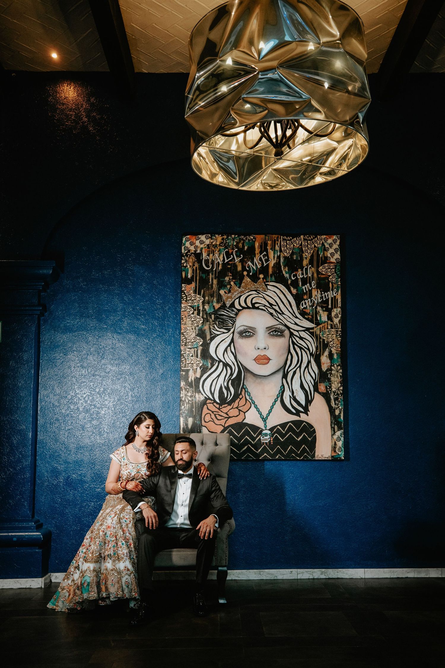Couple poses in front of blue wall with artistic portrait and decorative chandelier in moody lighting.