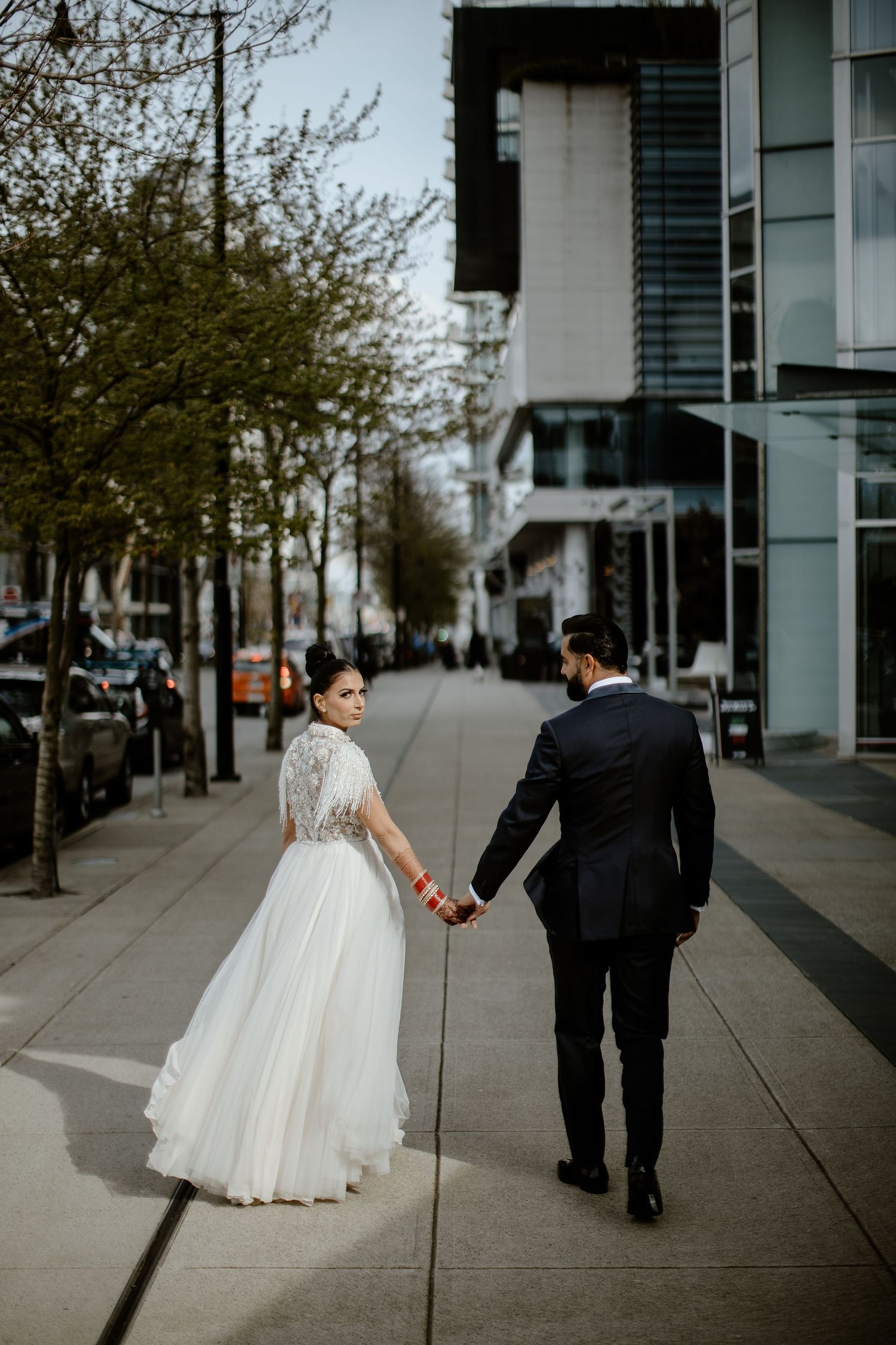 A couple in wedding attire walks along a modern urban sidewalk with glass buildings and trees in the background.