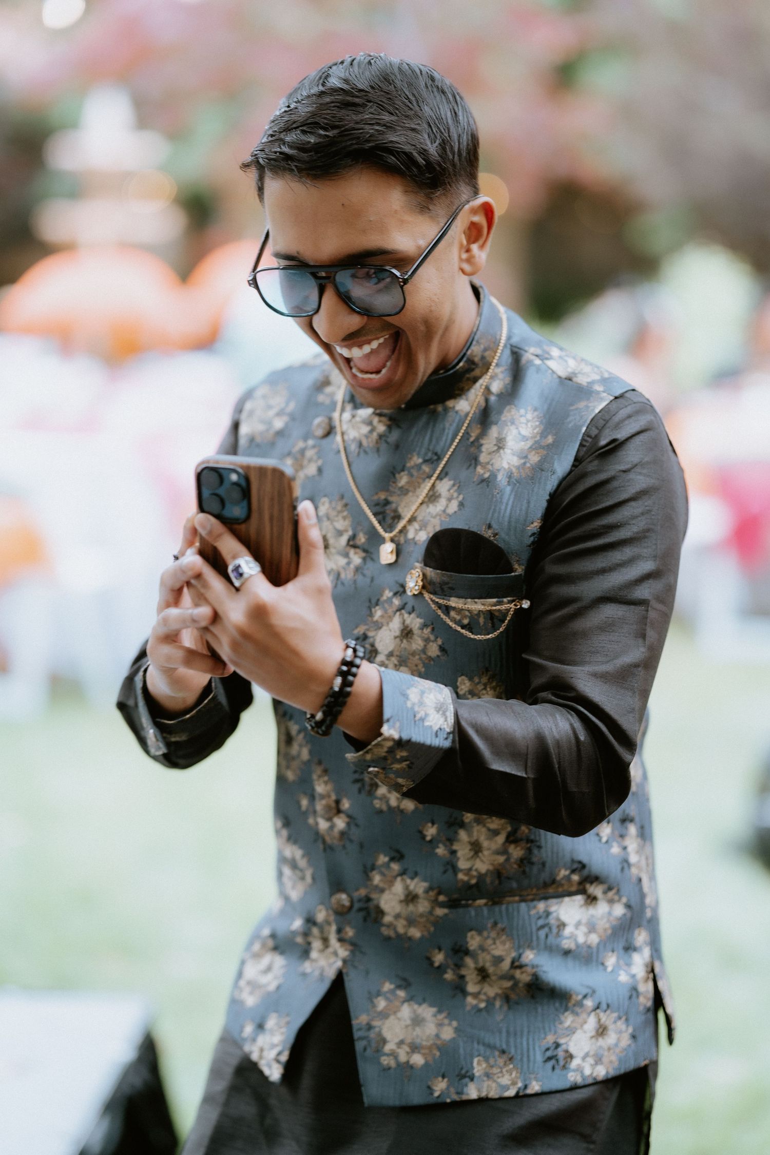 A person wearing glasses and a floral patterned jacket smiles while looking at their mobile phone outdoors.