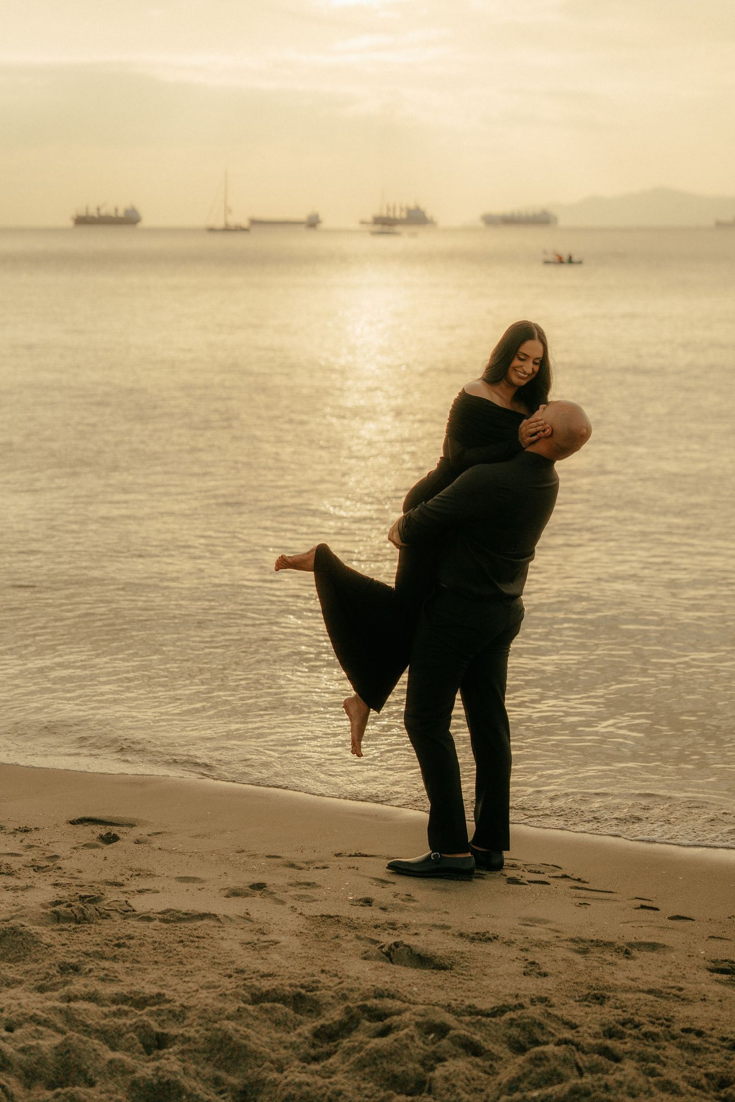 A series of silhouettes dance and play on a misty beach at sunset with ships visible on the horizon.