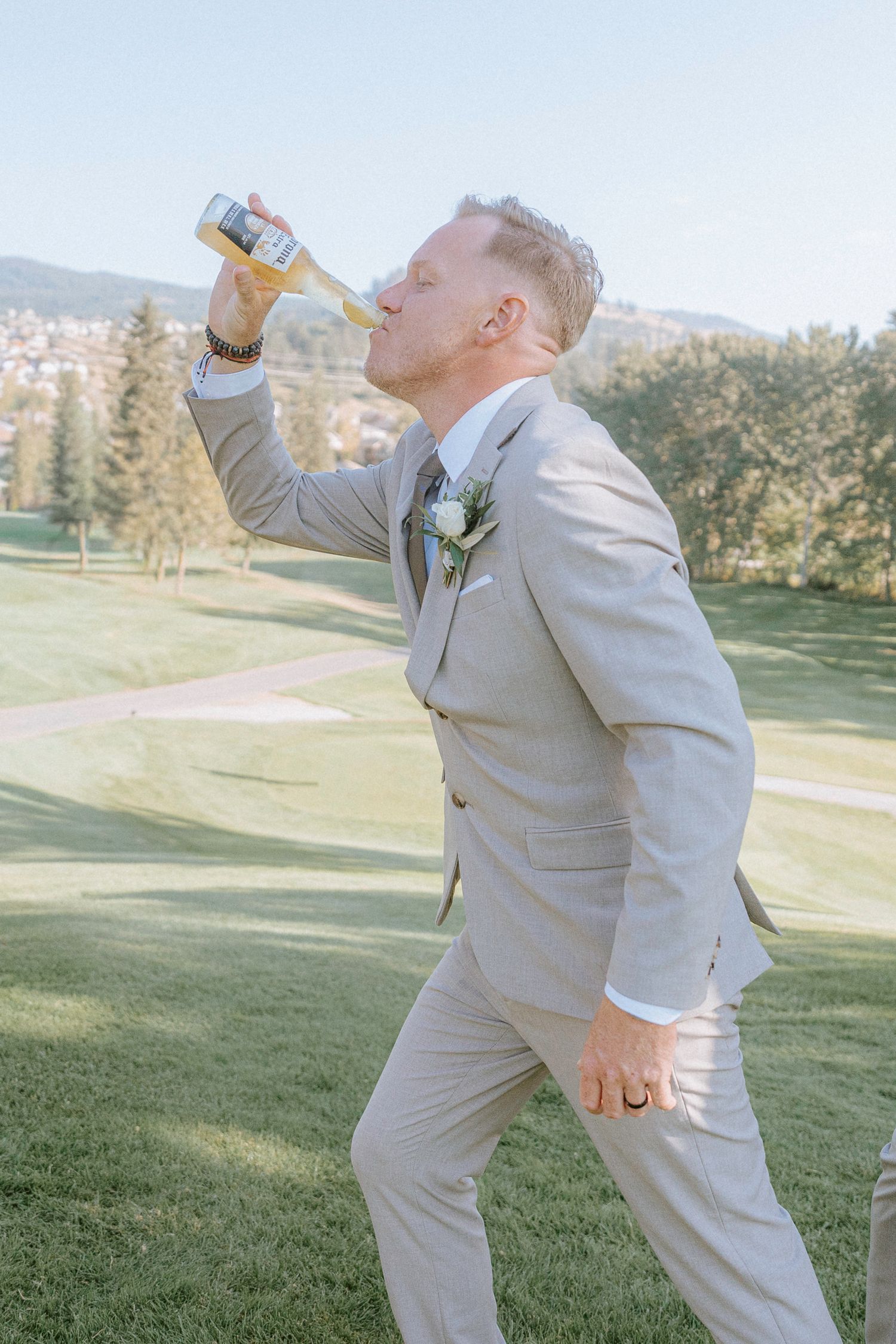Person in light gray suit strikes playful poses while drinking from a bottle on grassy hill.
