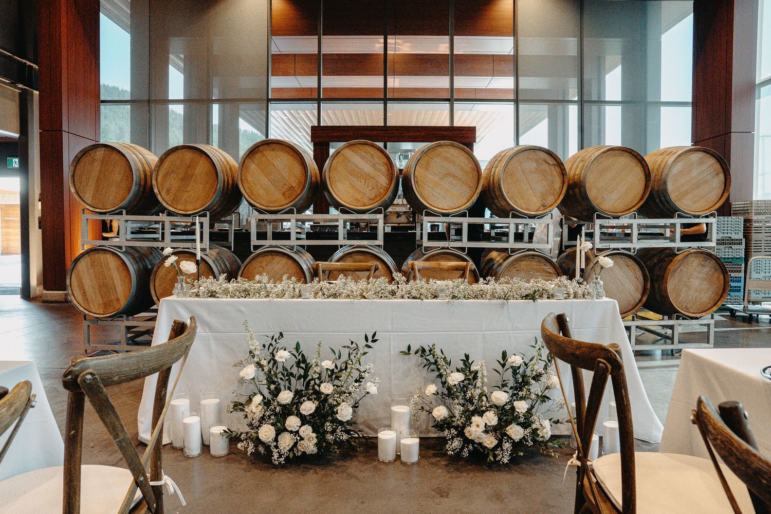 A modern winery event space with wooden barrels lining the wall and elegant white floral arrangements on display.