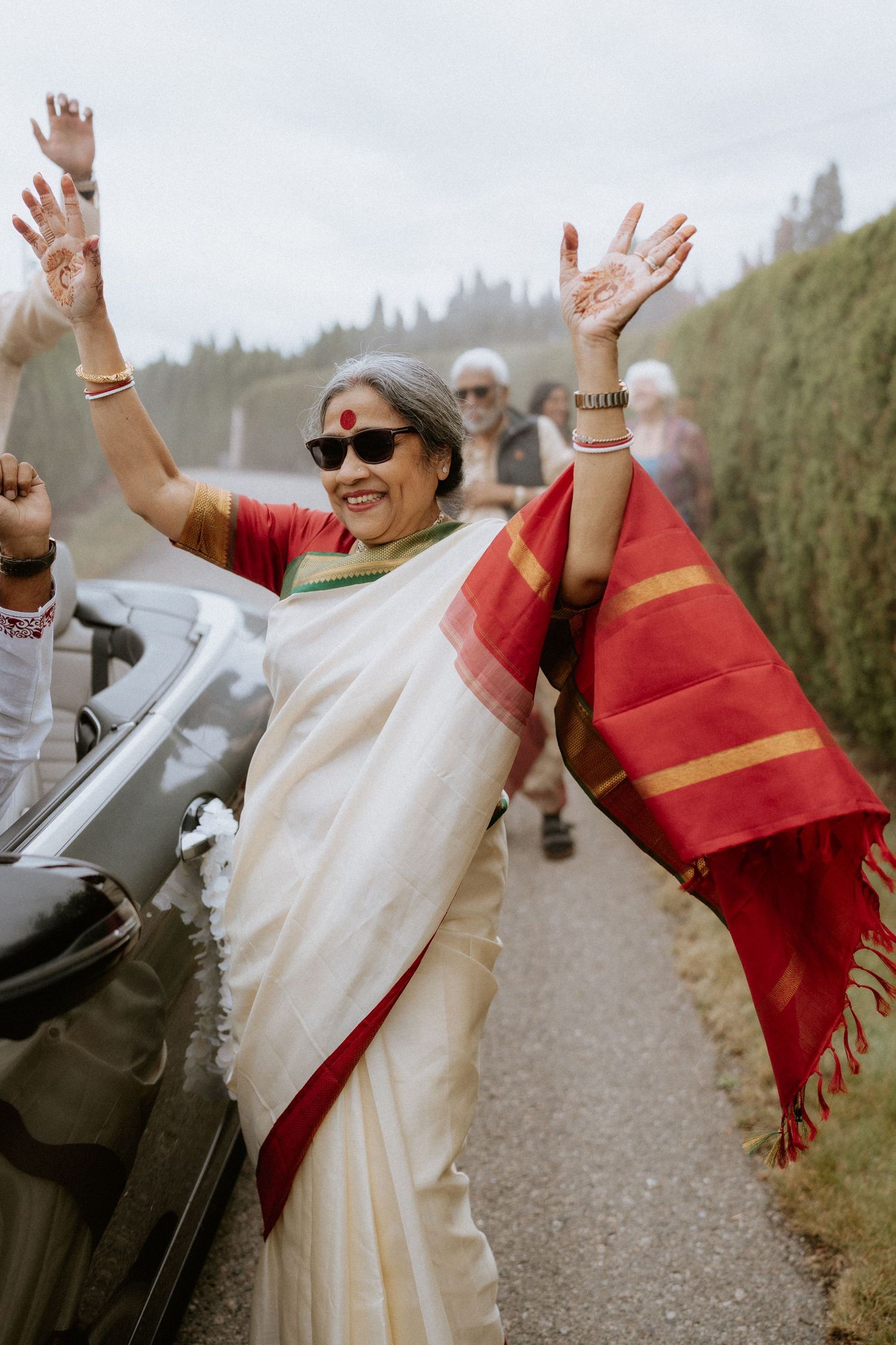 People celebrating in traditional Indian attire with raised arms on a pathway surrounded by greenery.