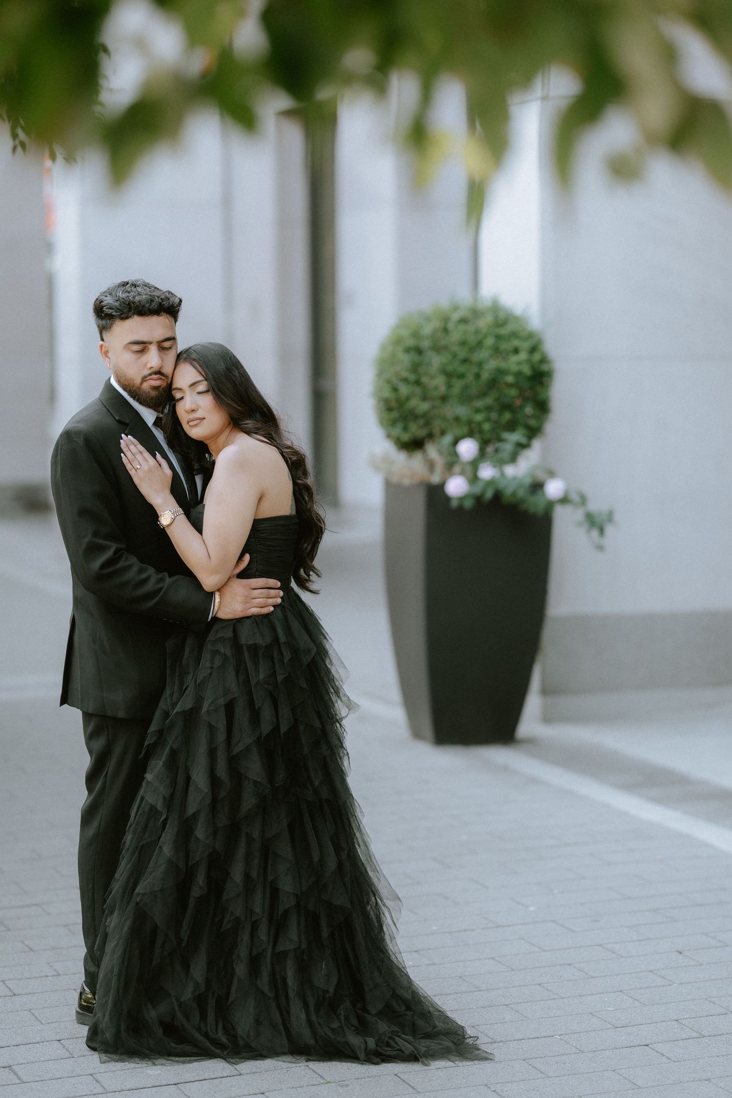 A romantic couple in formal black attire shares intimate moments during an outdoor photoshoot on a paved walkway.