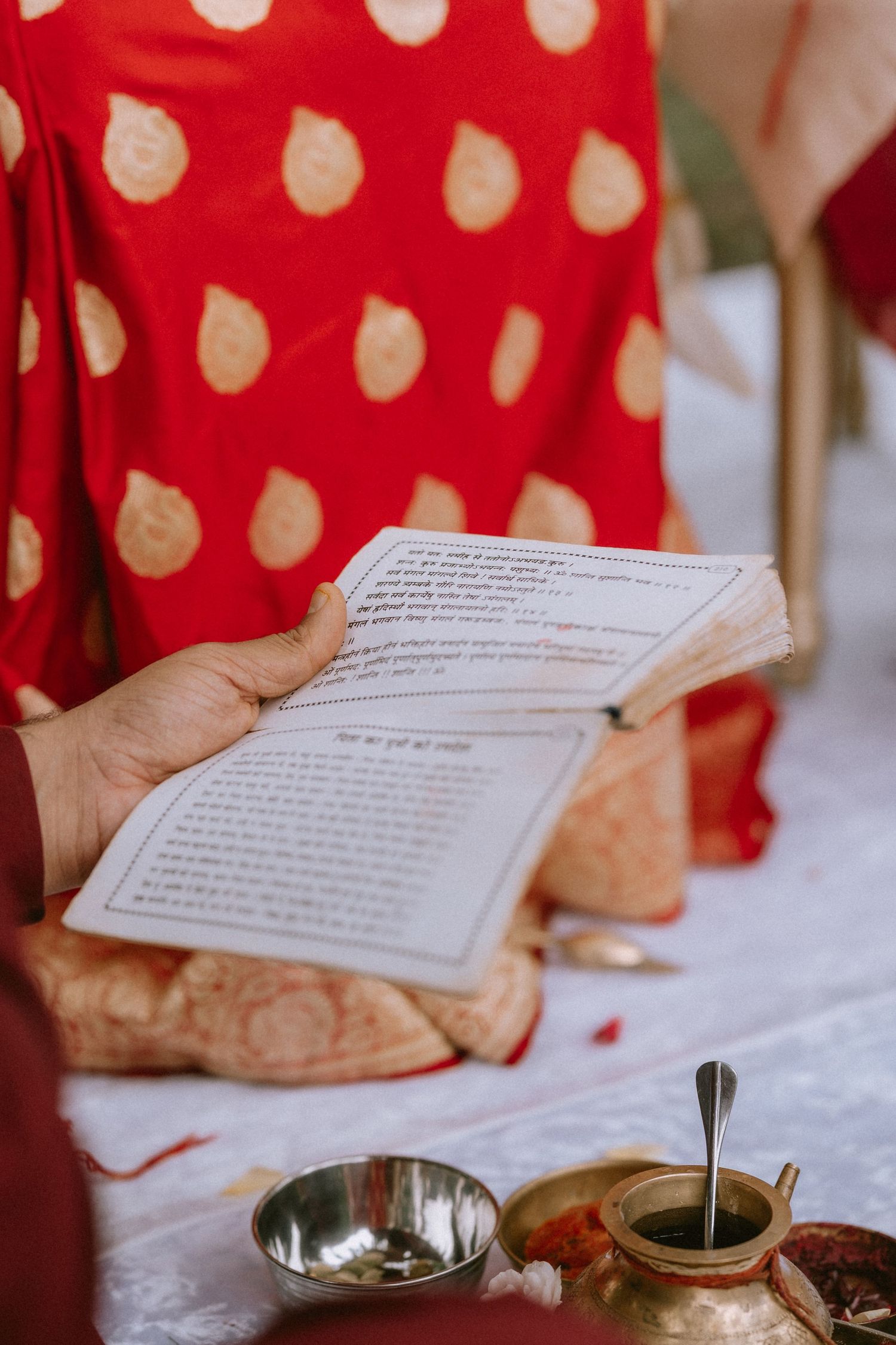 Hands holding ceremonial text with traditional mehndi designs against red polka dot fabric.