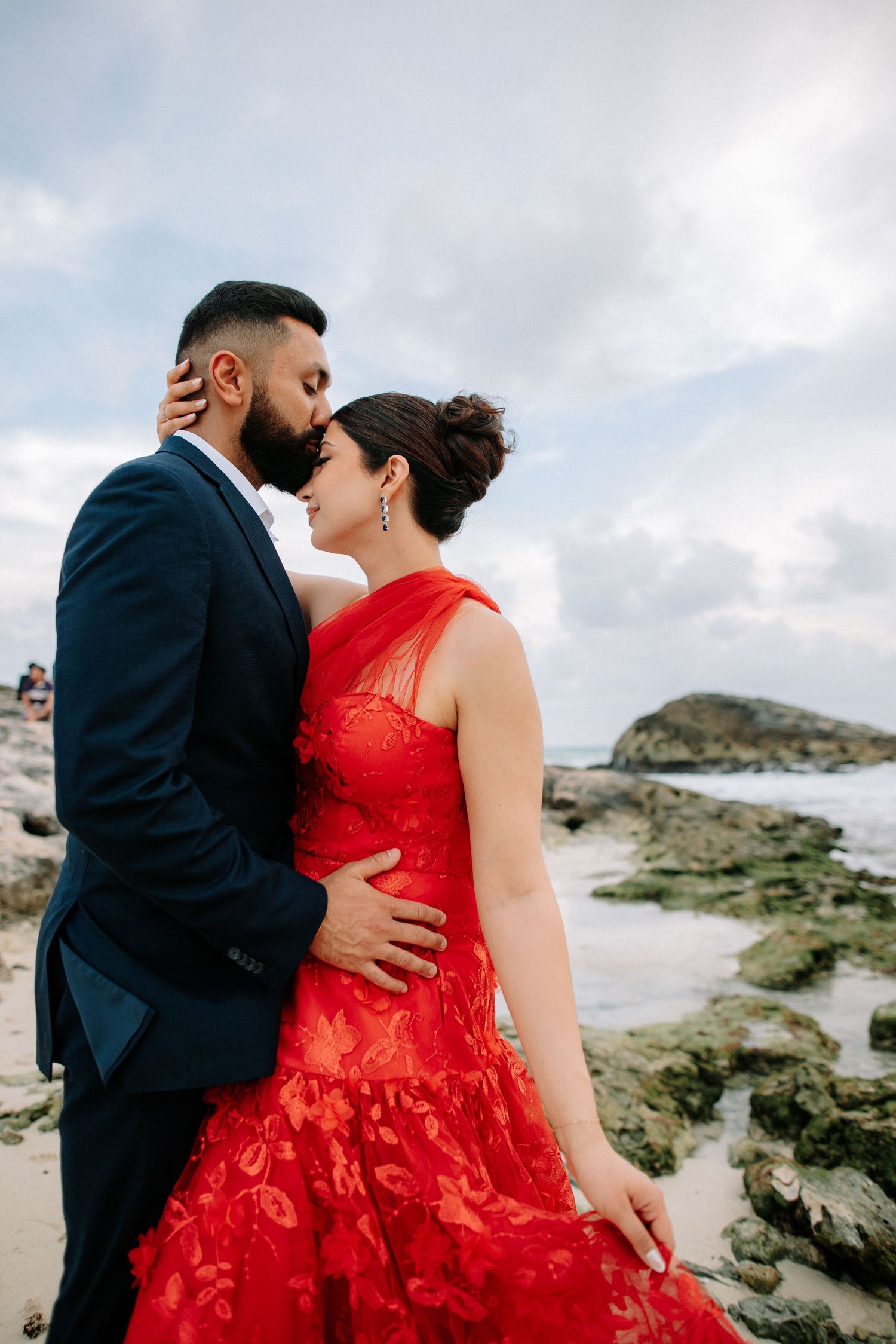 A couple in formal attire share intimate moments on a rocky beach, with the woman wearing a striking red lace dress.