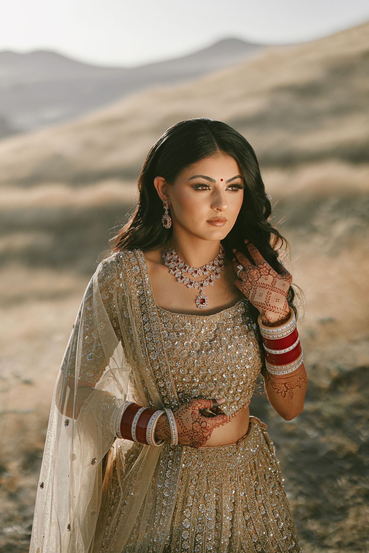 Traditional Indian bride wearing sparkly golden lehenga and jewelry poses in desert mountain landscape at sunset.