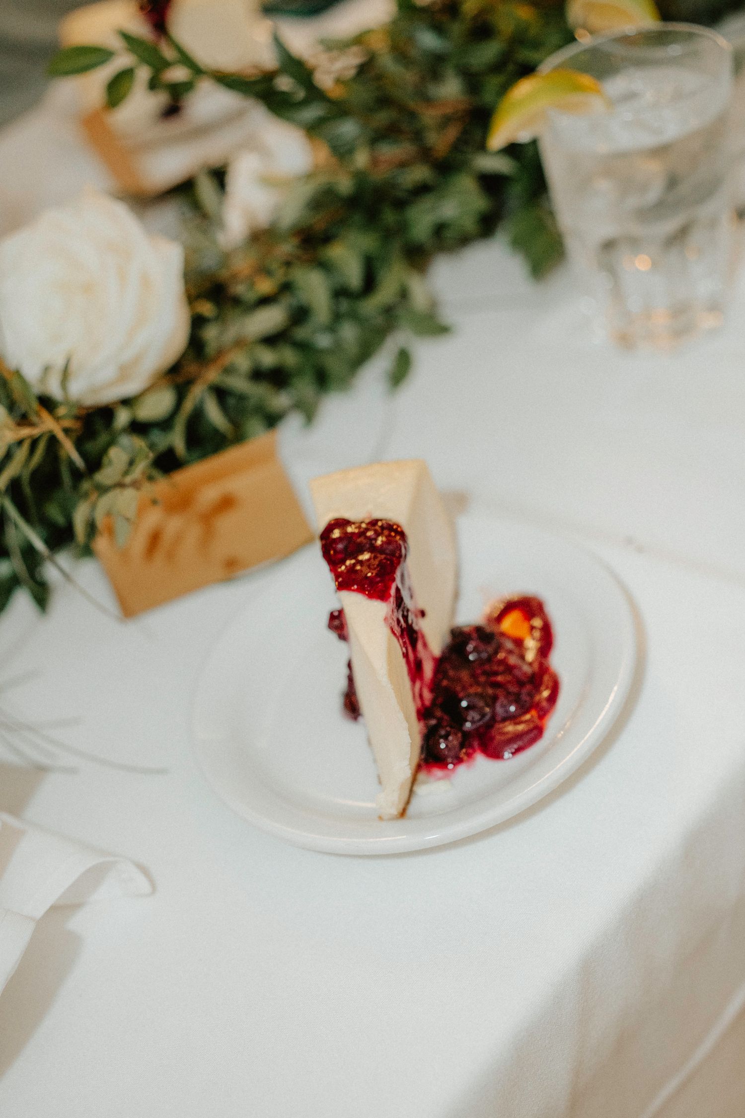 Slices of red berry cheesecake on white plates with greenery decorations on a tablecloth.