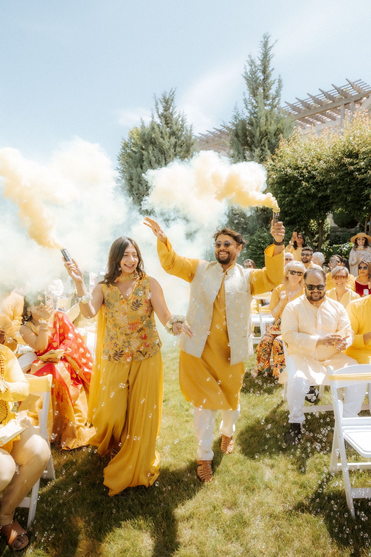 A group celebrates outdoors with yellow smoke bombs and coordinated yellow and white outfits in a garden setting.