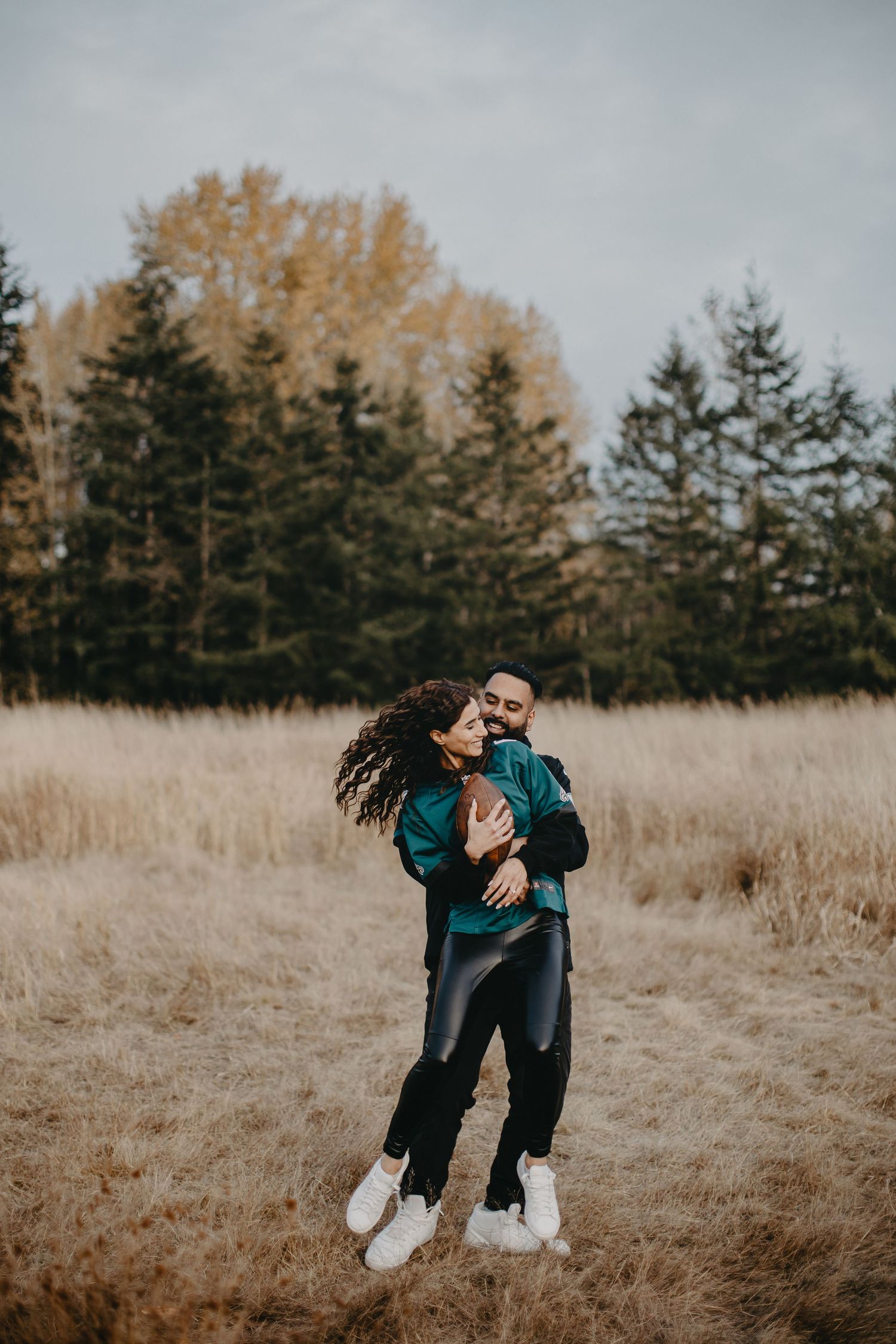 Couple in coordinated teal outfits embrace playfully in a golden autumn field with evergreen trees in background.