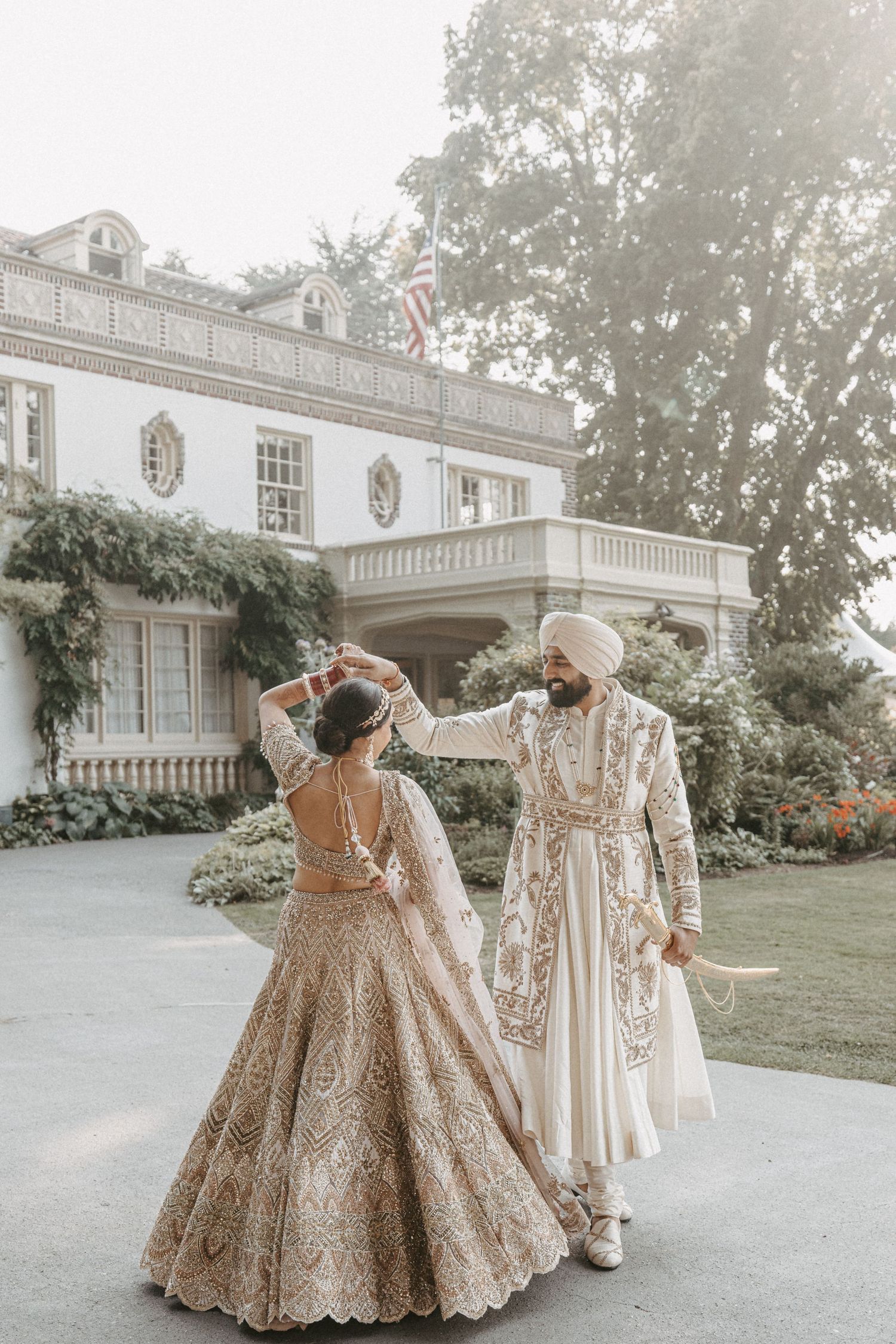 A couple in traditional Indian wedding attire dances together outside an elegant white mansion.