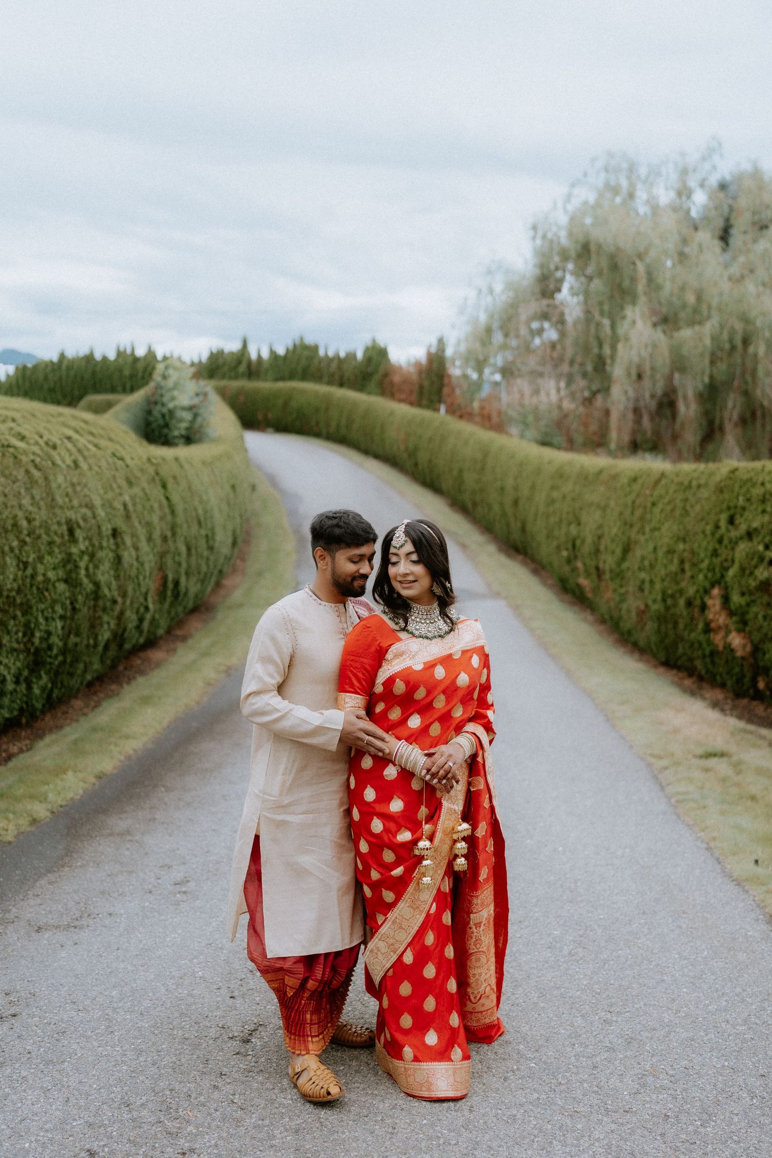 A romantic couple walks along a path lined with tall manicured hedges on a cloudy day.