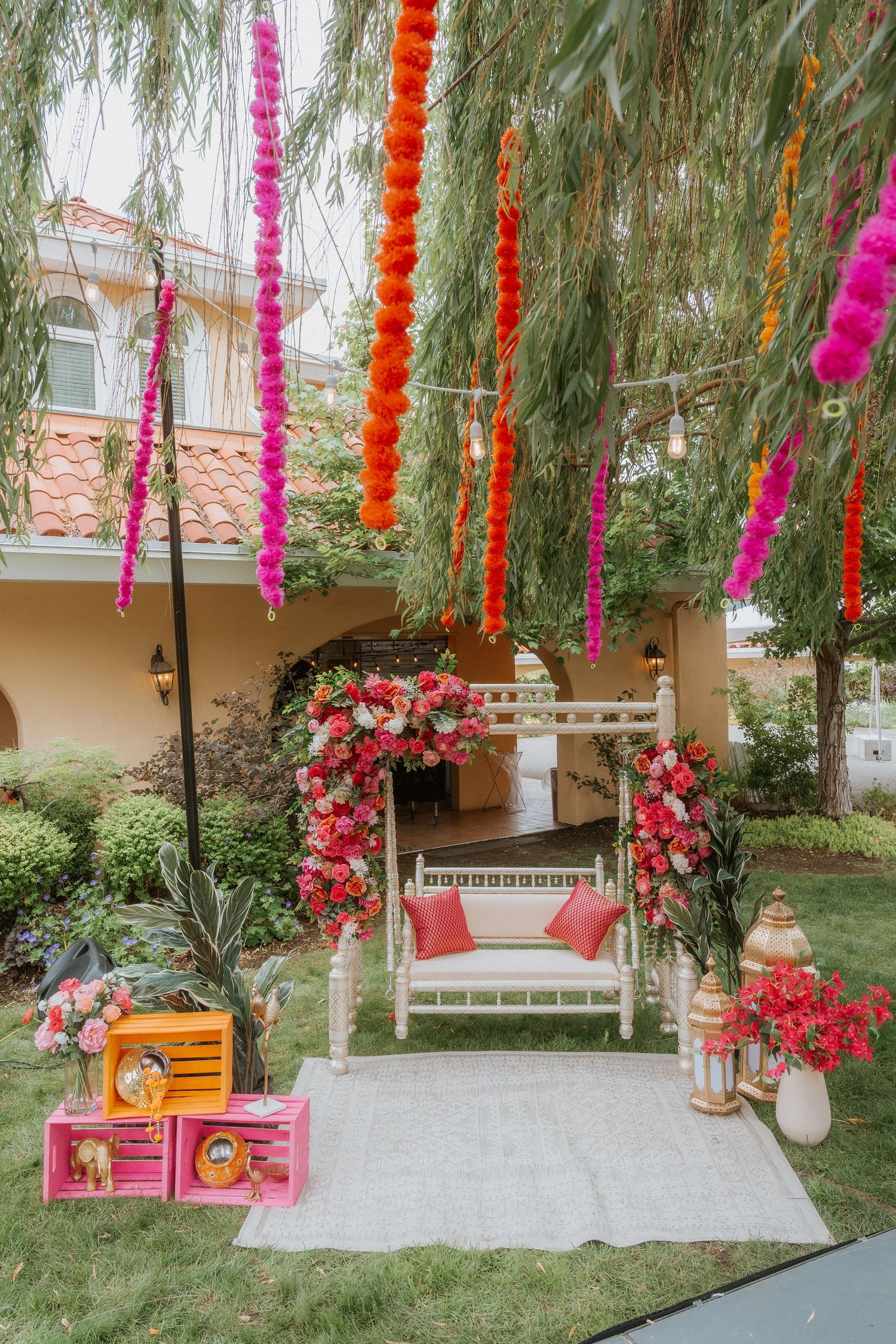 Colorful wedding decoration setup with pink and orange streamers hanging from trees over a white bench with floral arch.