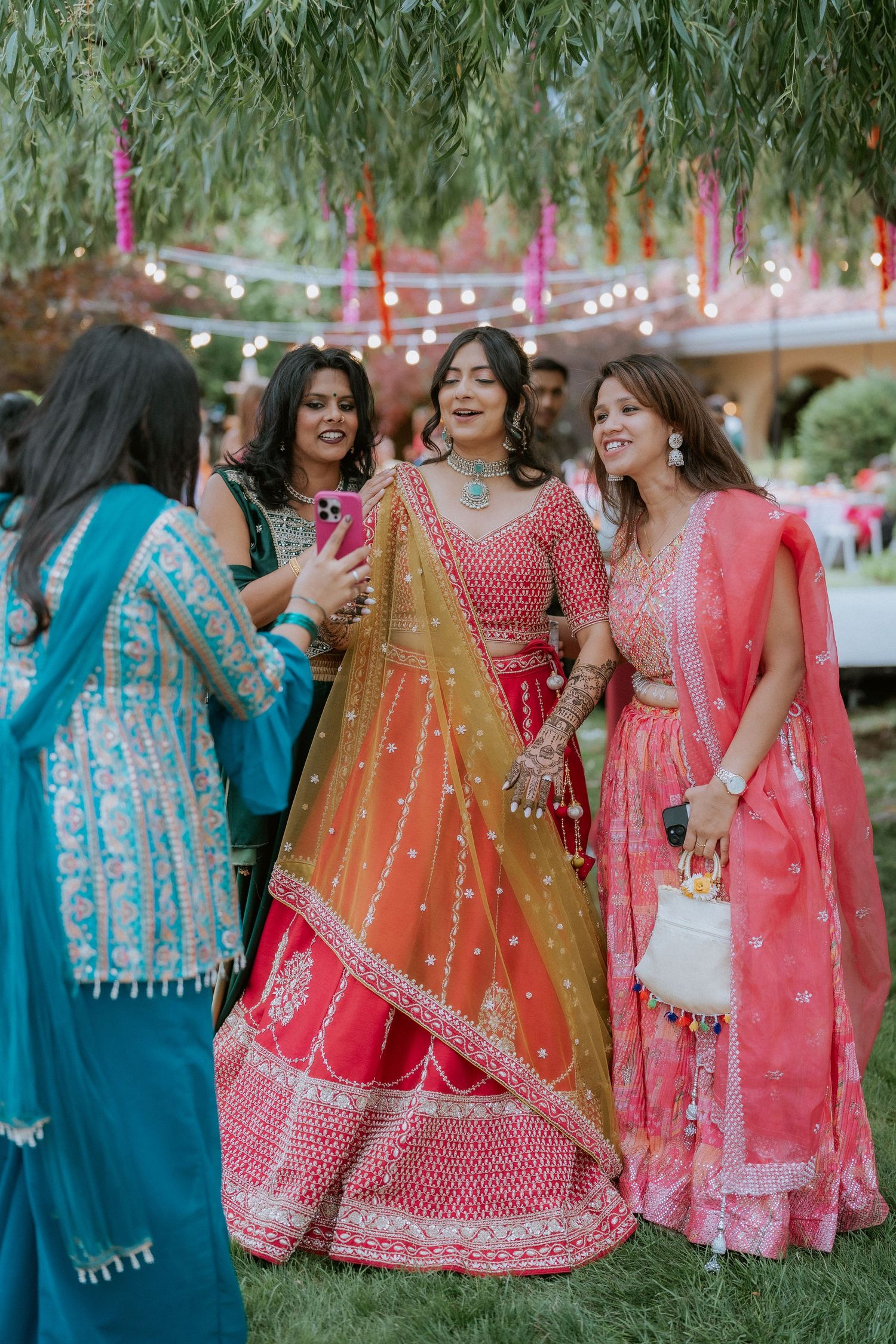 Women in colorful traditional Indian lehengas and sarees laugh together at an outdoor celebration.