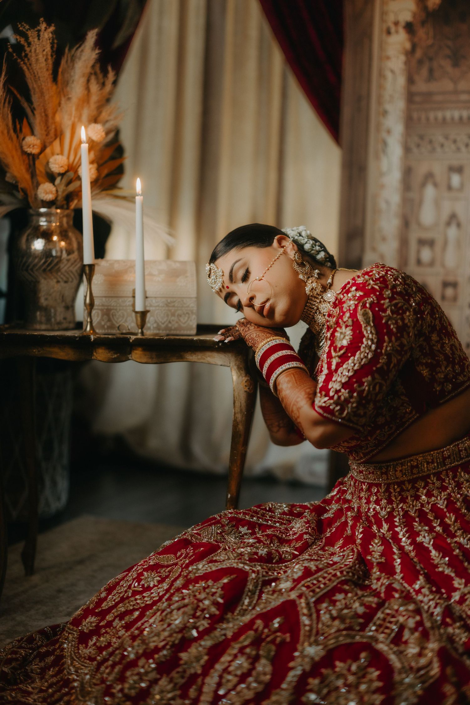 A traditional Indian bride in red lehenga poses gracefully with candlelight in an ornate interior setting.