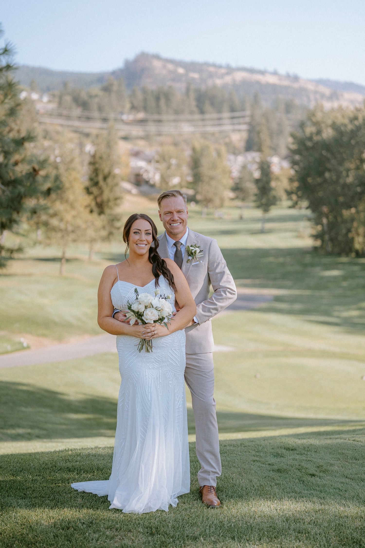 A bride and groom embrace on rolling green hills of a golf course with mountains in the background.