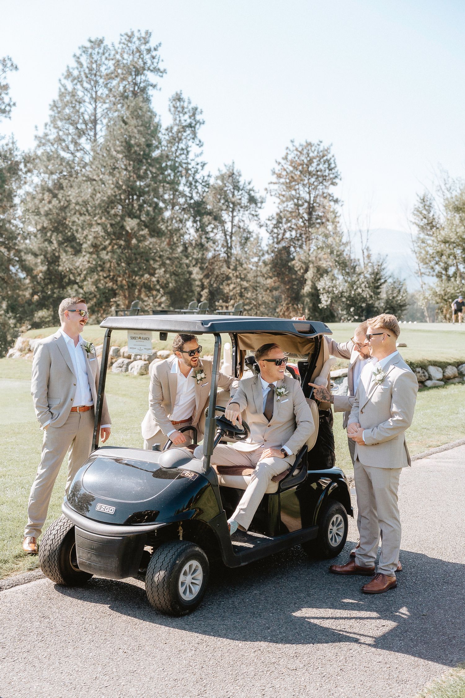 Groomsmen in light gray suits gather around a golf cart on a sunny day at a wedding venue.