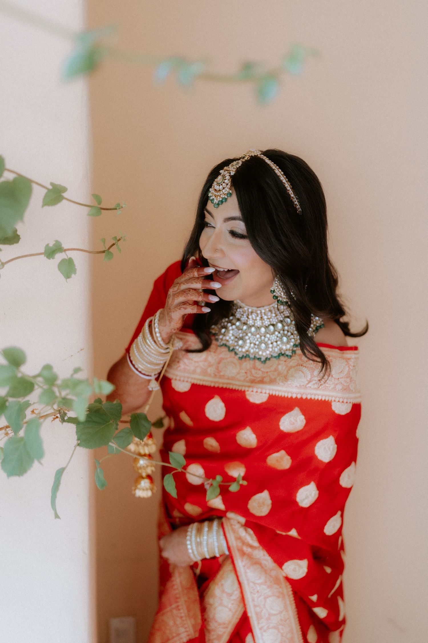 Bride in traditional red and gold polka dot sari poses gracefully with flowing fabric in indoor setting.