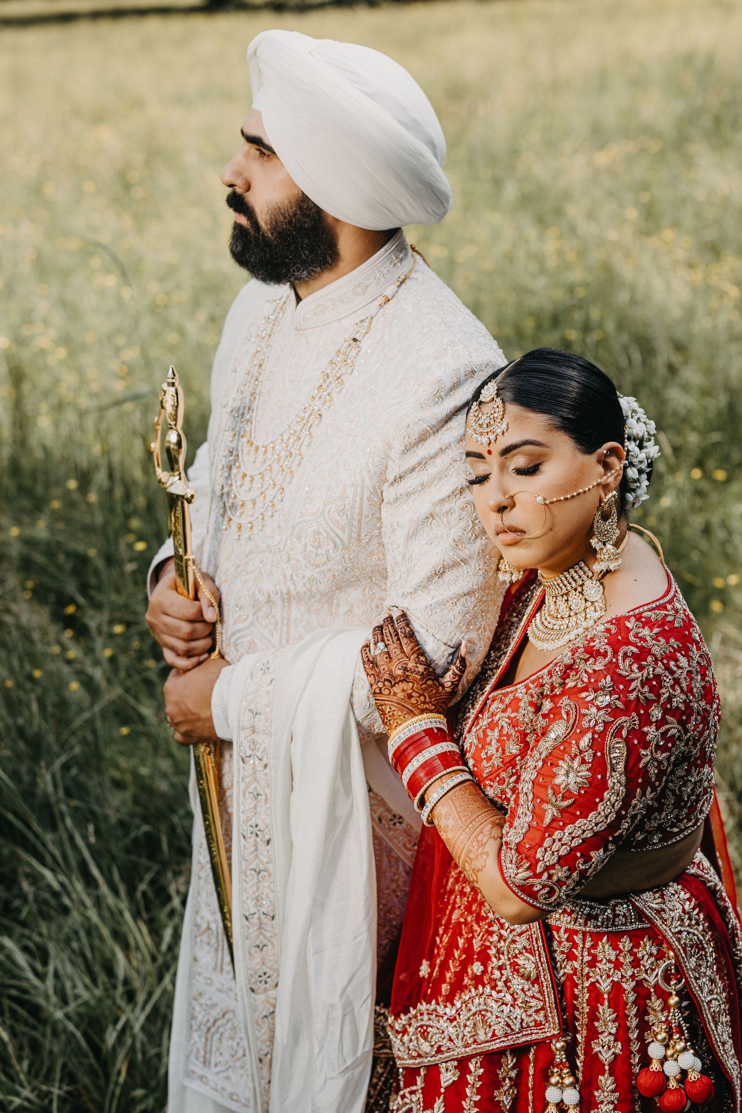 Traditional Indian wedding couple in elegant attire poses outdoors, with bride in red lehenga and groom in white sherwani.
