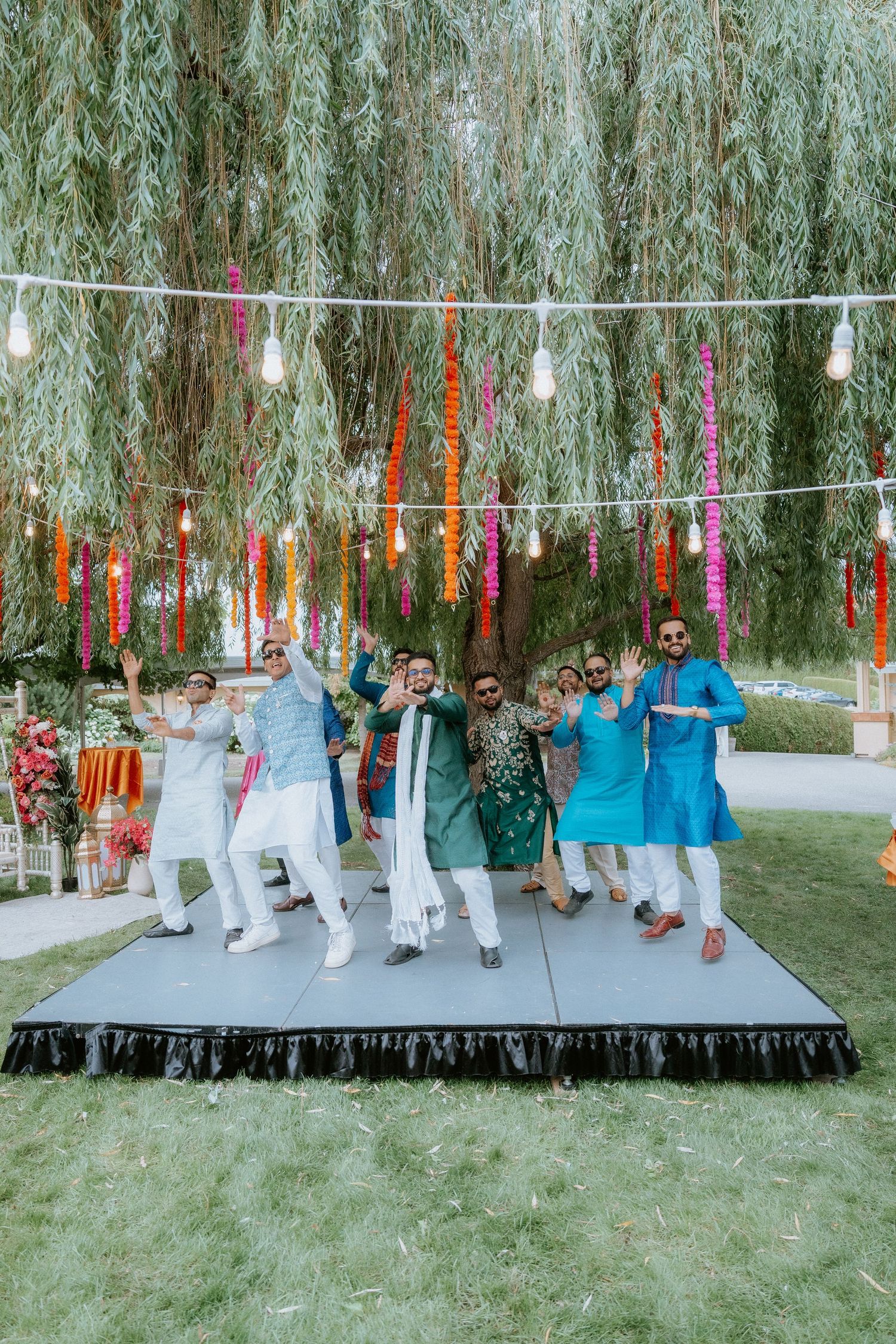 Wedding party poses under a weeping willow tree with colorful ribbons and string lights creating a festive outdoor atmosphere.