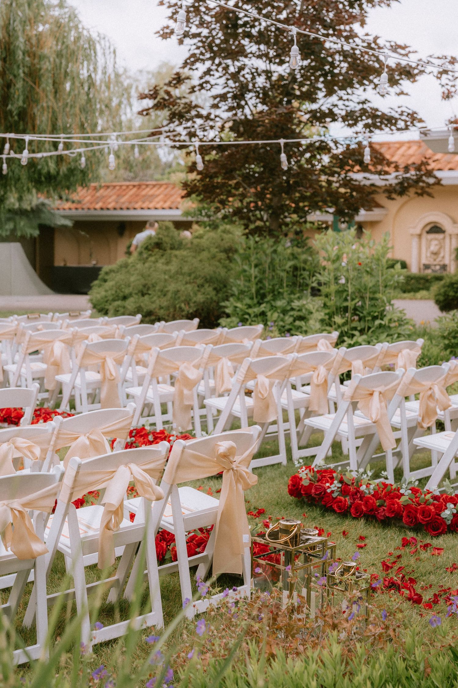 White wedding chairs arranged outdoors with red floral decorations along the aisle.