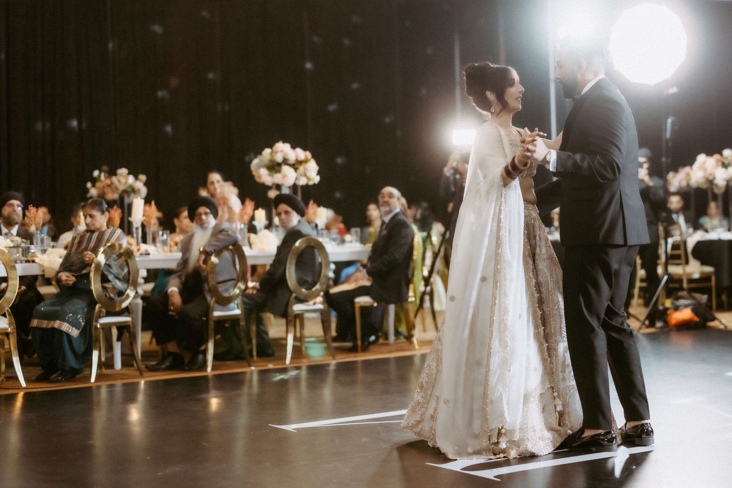 A couple shares their first dance at a wedding reception under dramatic lighting while guests watch from their seats.