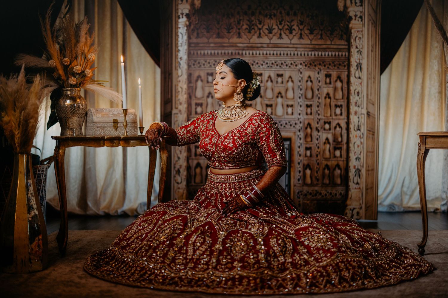 Bride in ornate red lehenga poses elegantly on floor in traditional Indian wedding attire against textured wall backdrop.