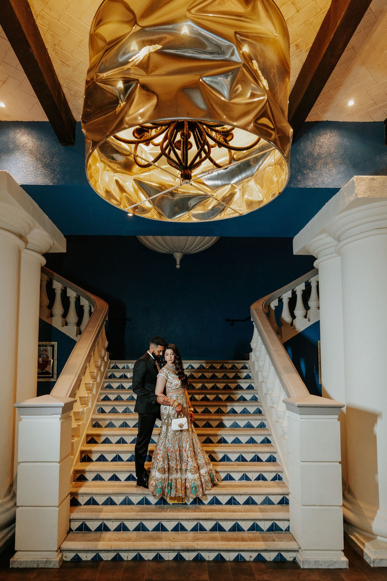 Couple stands on geometric patterned staircase under golden light fixtures in elegant venue interior.