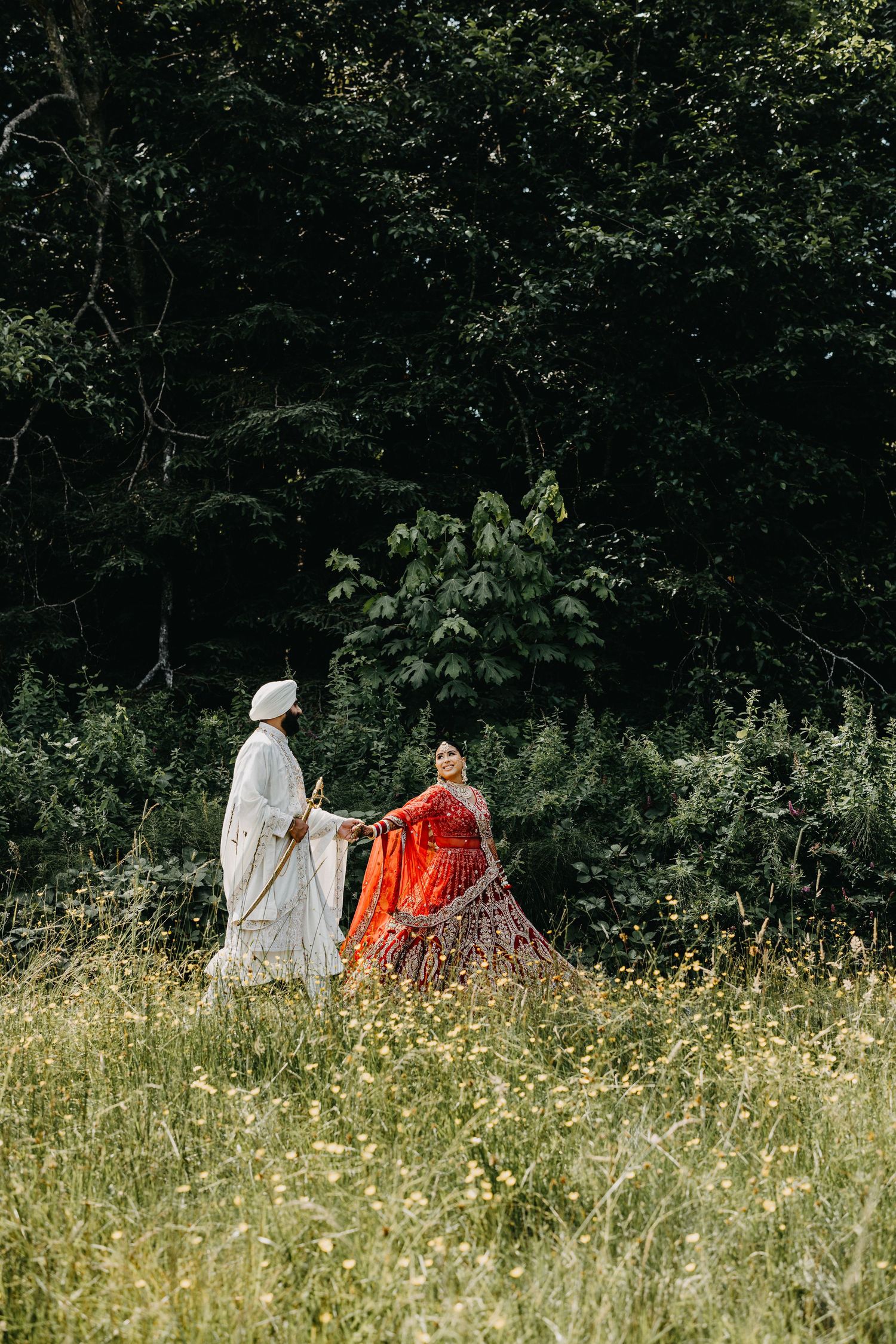 Two figures in period costumes, one in white and one in red, sit together in a wildflower meadow against a dark forest backdrop.
