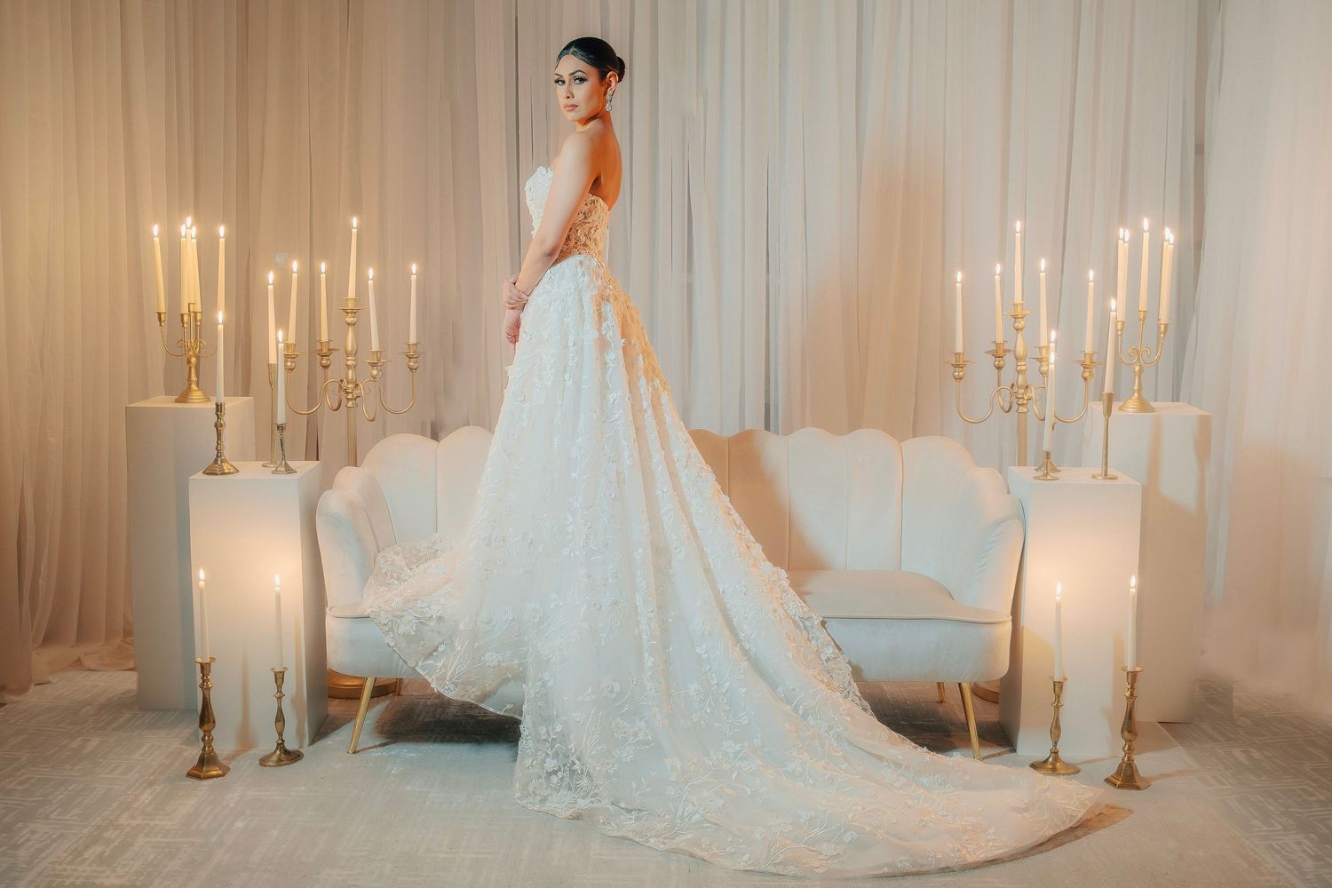 Bride in flowing white lace wedding gown stands elegantly among tall white candles in an ethereal setting.