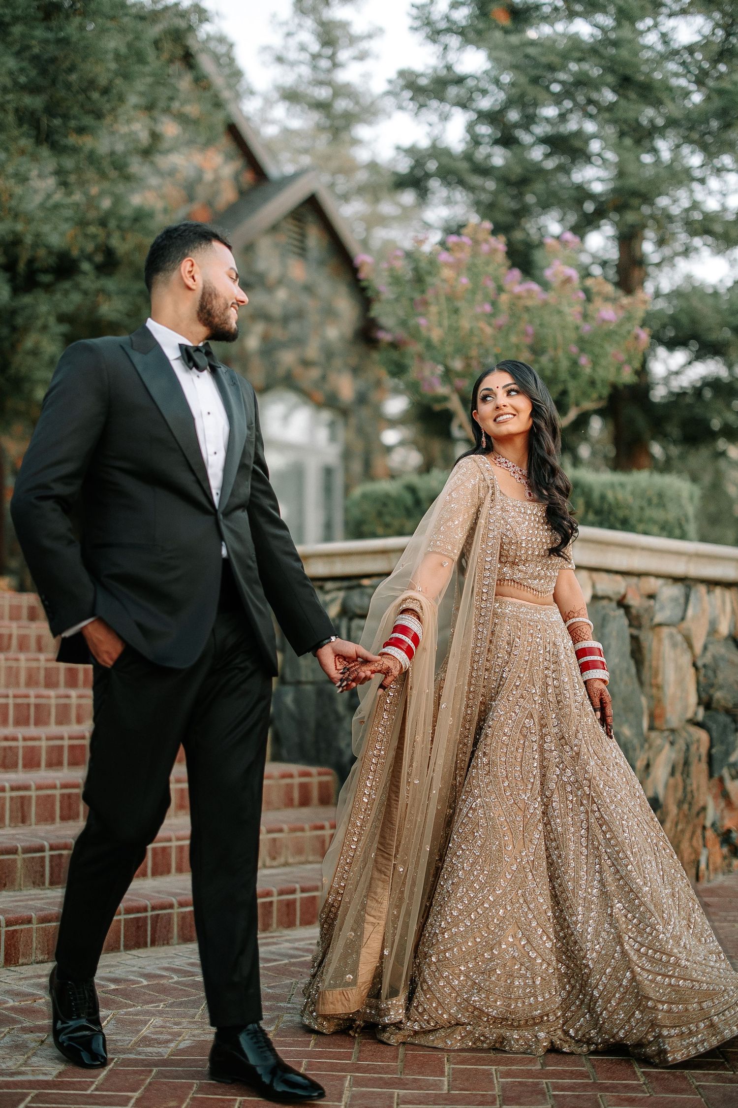 Couple in wedding attire with bride wearing traditional gold lehenga and groom in black suit standing on brick steps.