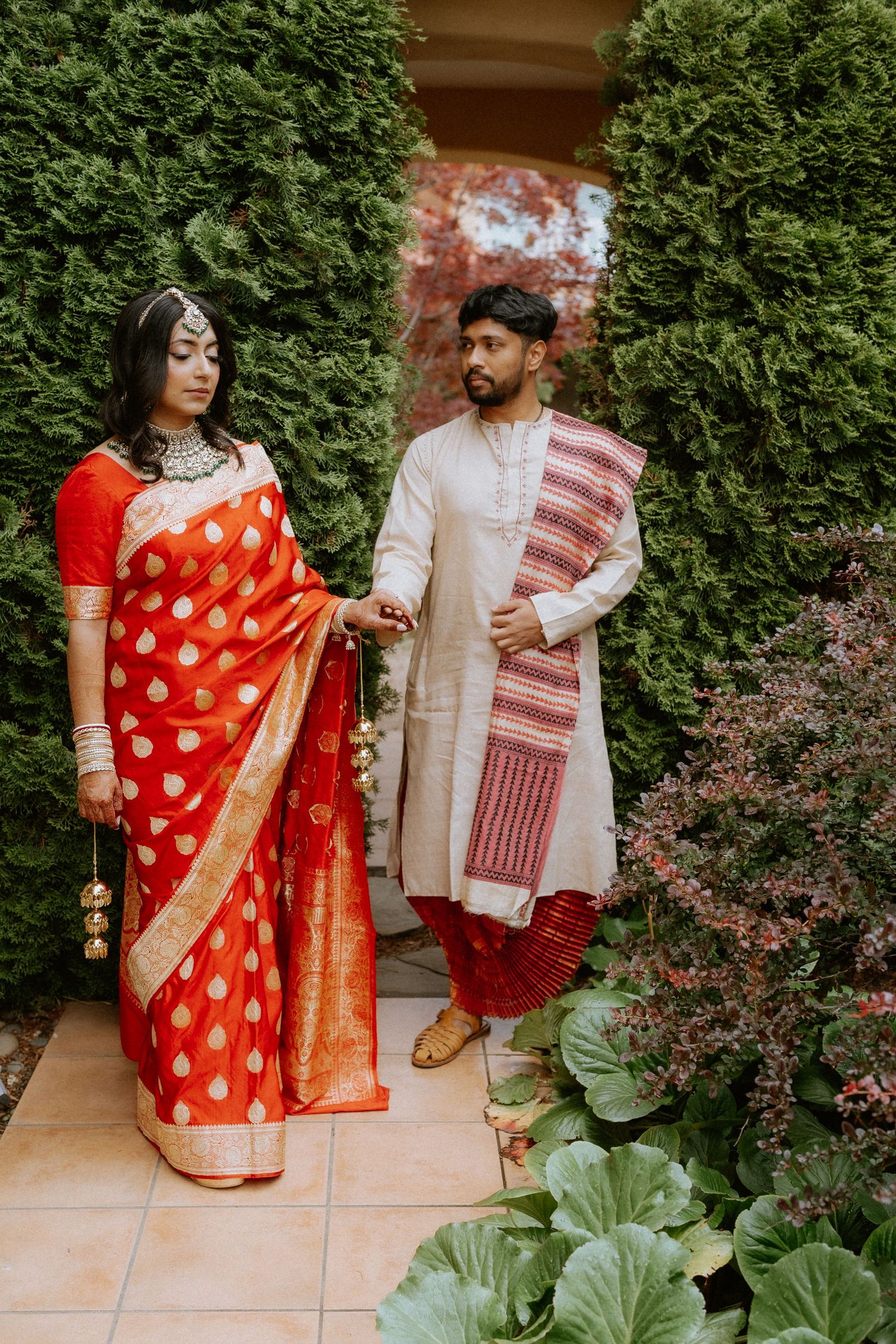 A couple in traditional Indian attire poses in a garden setting with lush greenery and red flowers.