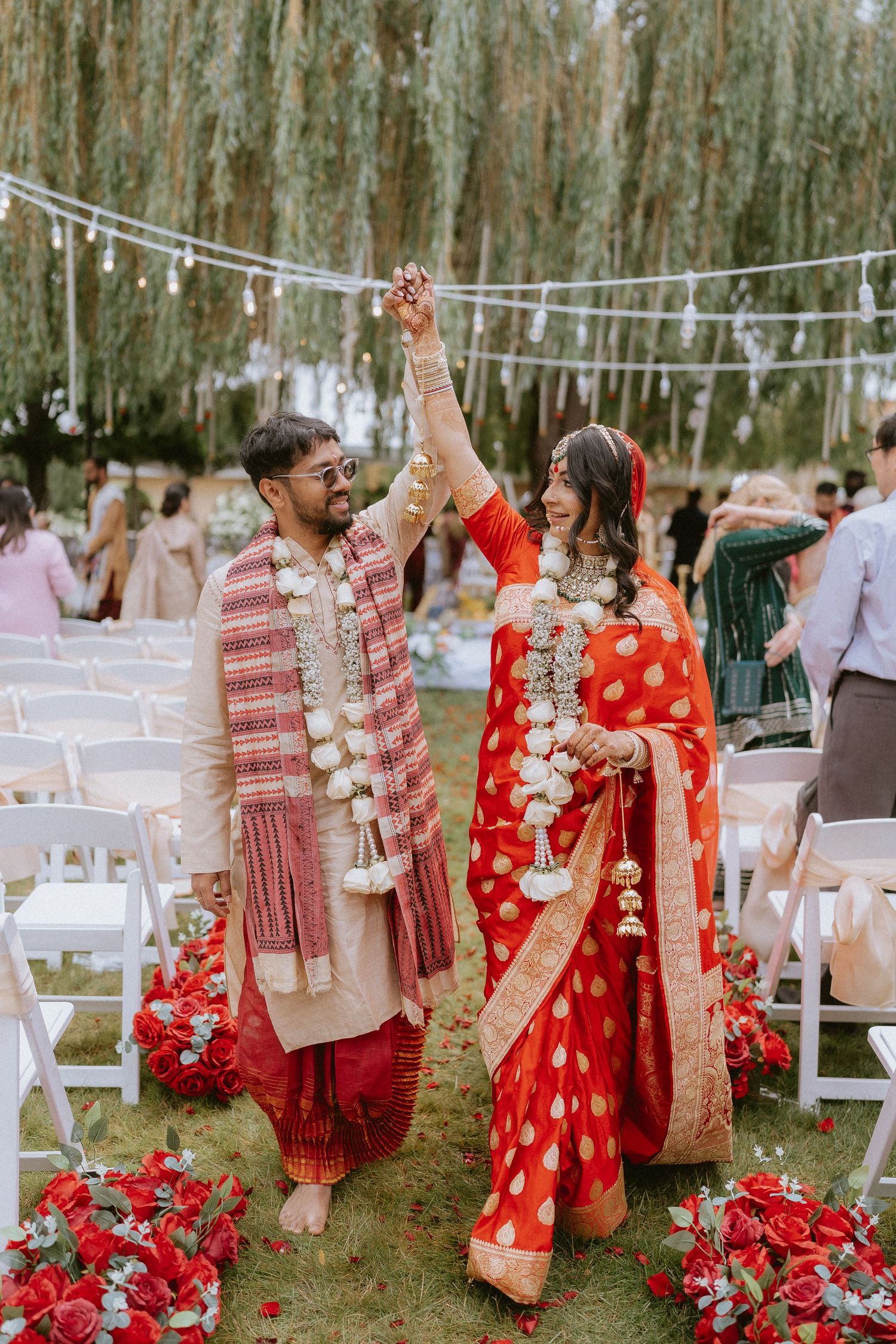 Outdoor wedding celebration with guests in red and white attire among trees with string lights and white chairs.