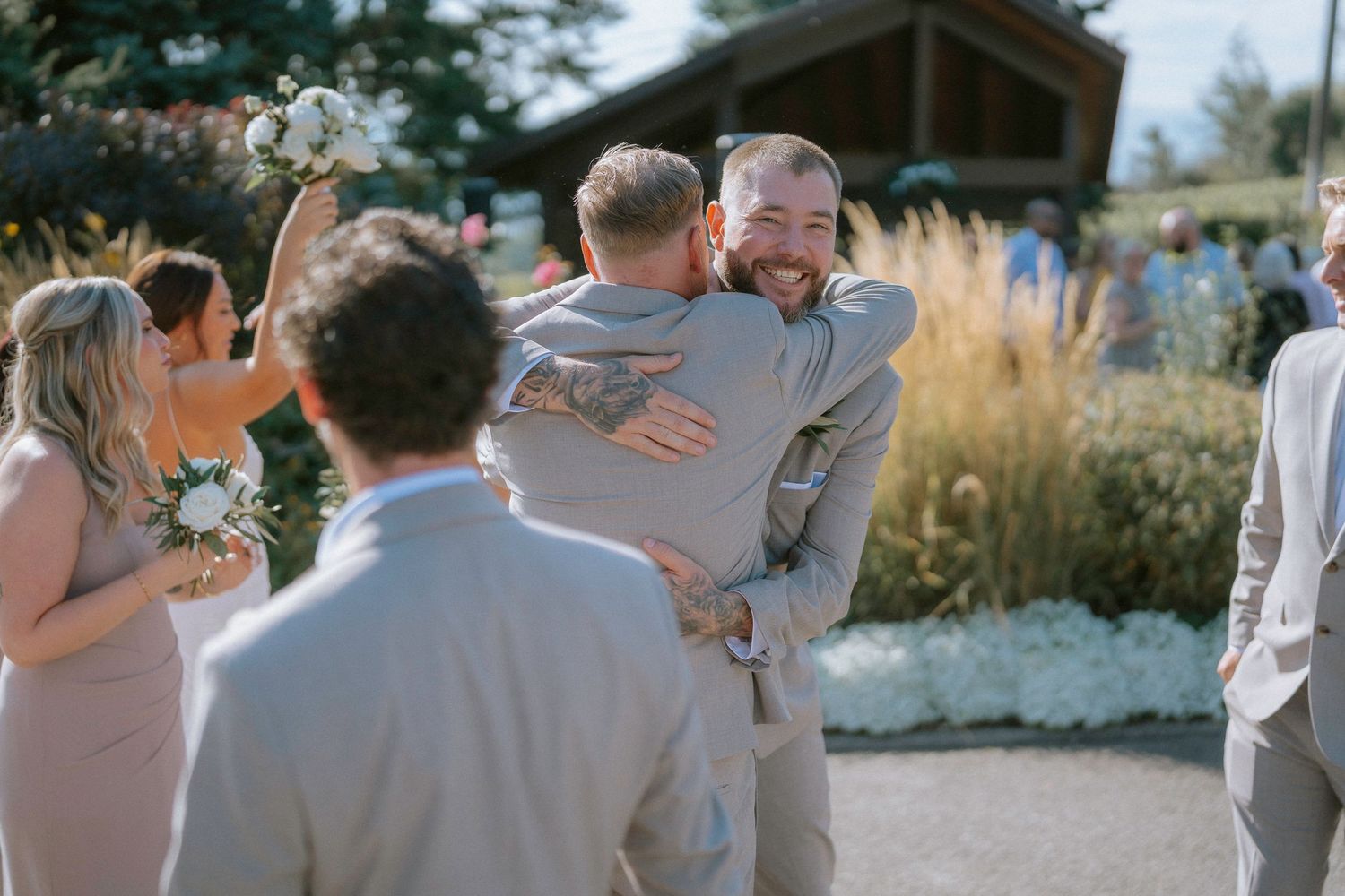 Wedding party members sharing joyful embraces during outdoor celebration with ornamental grasses in background.