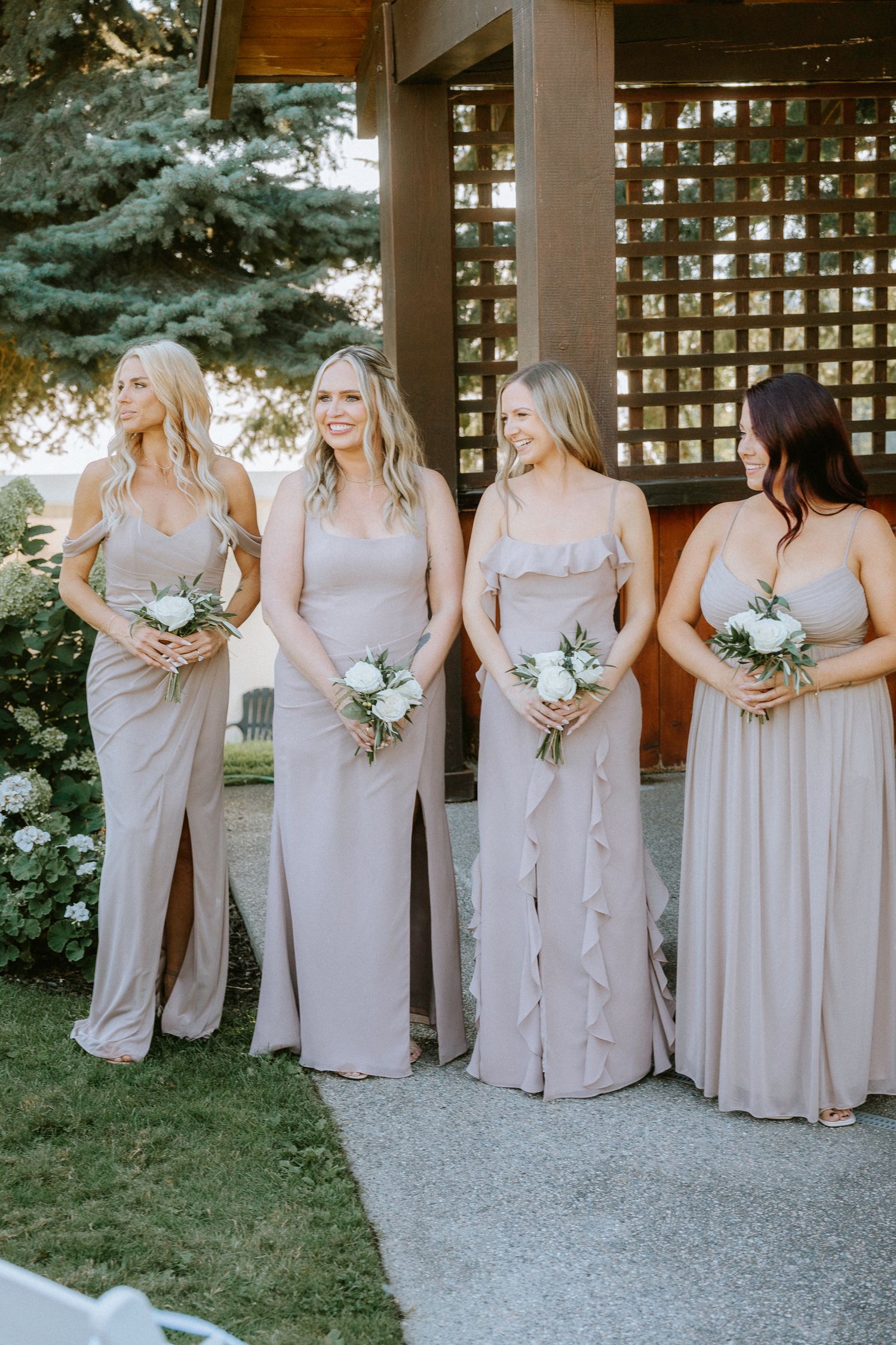 Bridesmaids in light pink dresses holding white bouquets pose together at an outdoor wedding venue.