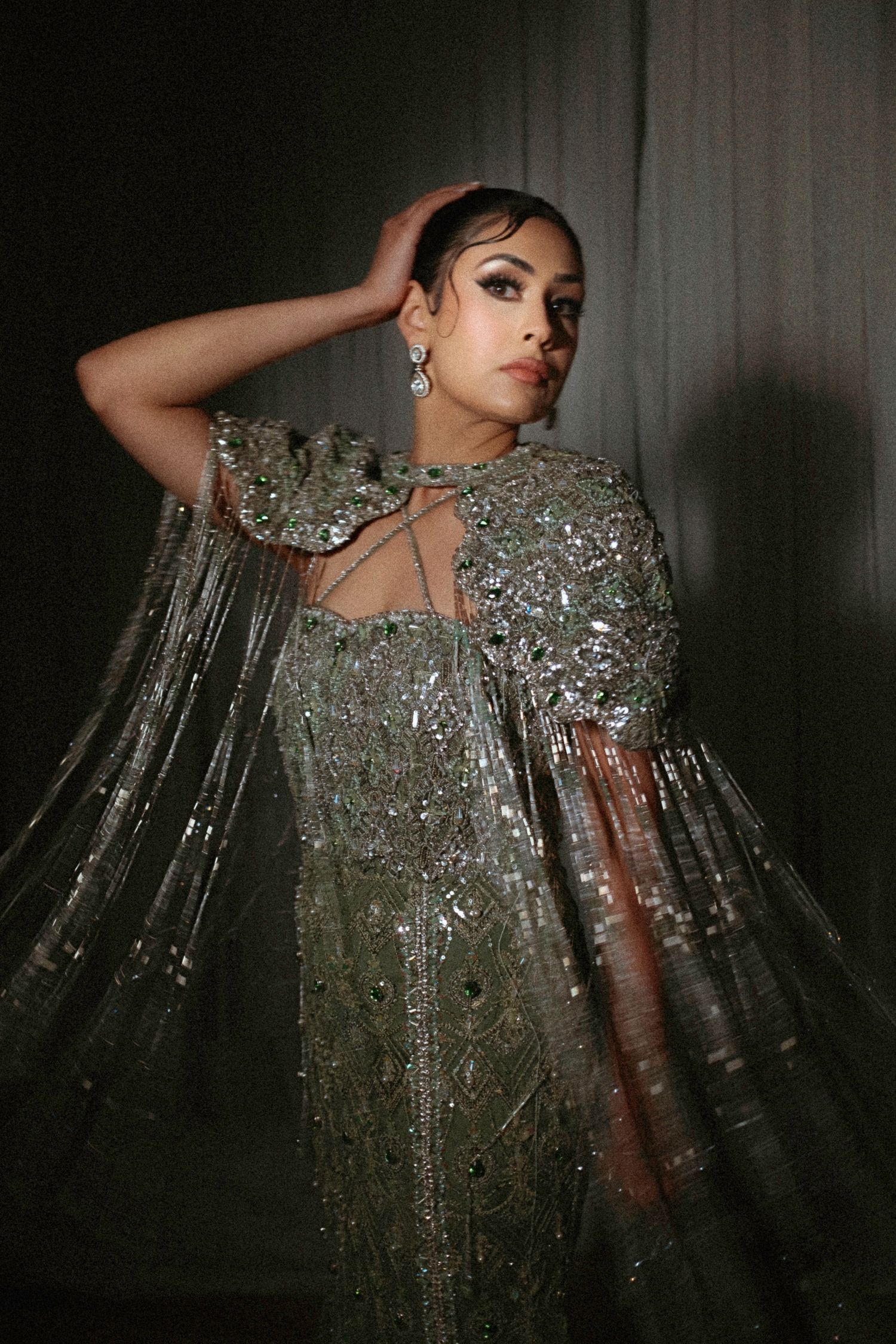 A model poses in a dramatic silver sequined gown with sheer cape sleeves against a dark background.