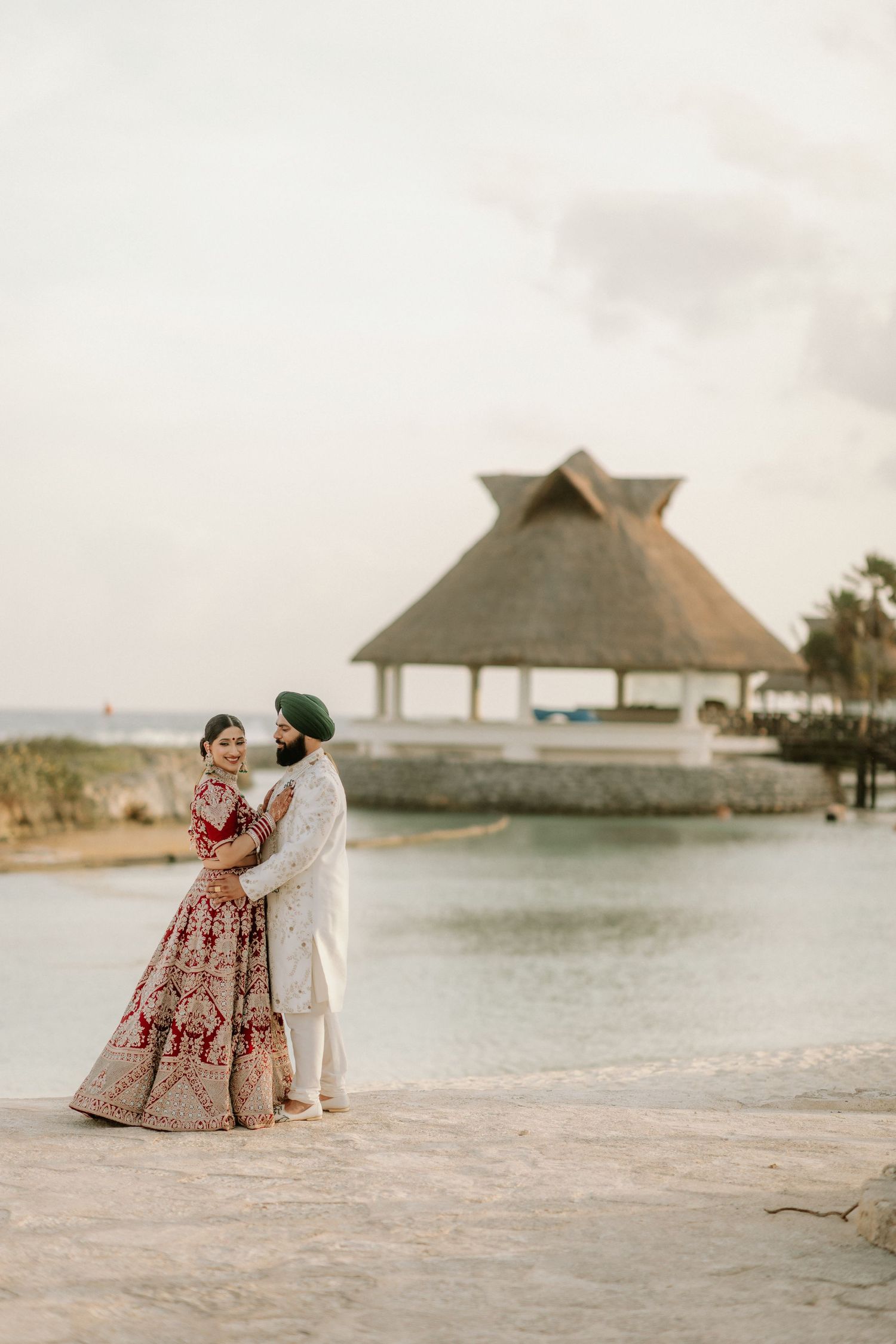 A couple in traditional Indian wedding attire embrace on a tropical beach with a thatched-roof gazebo in the background.