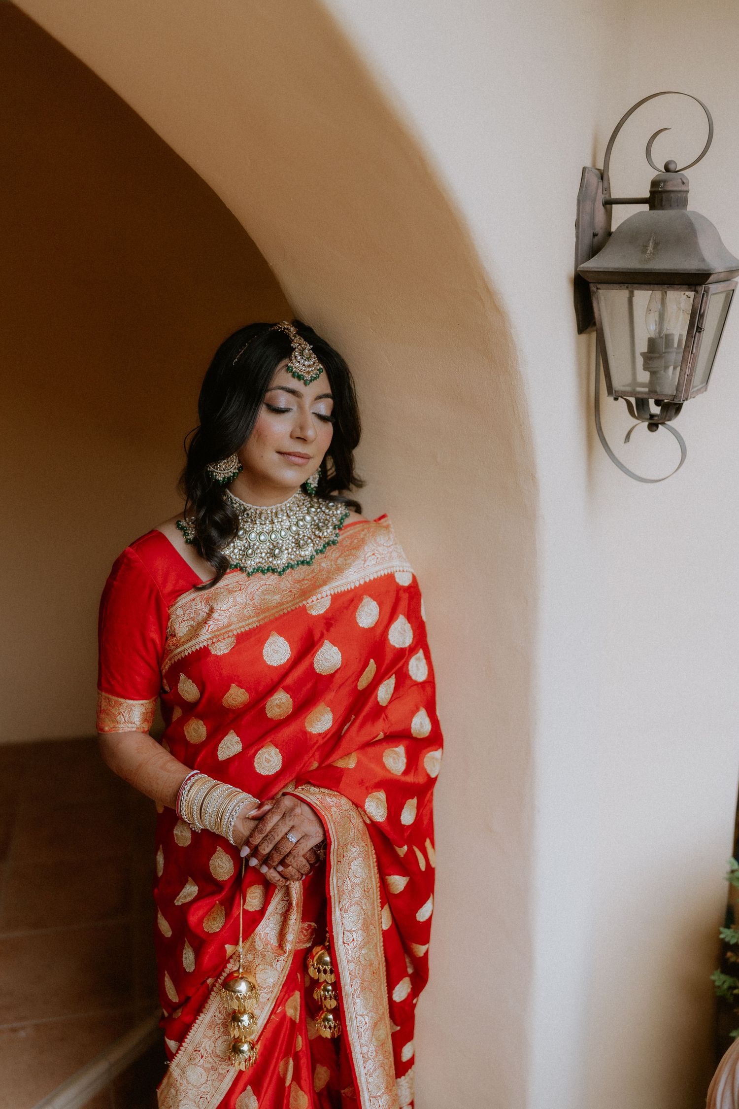 Indian bride in a vibrant red and gold saree poses elegantly against a white wall with decorative wall sconces.