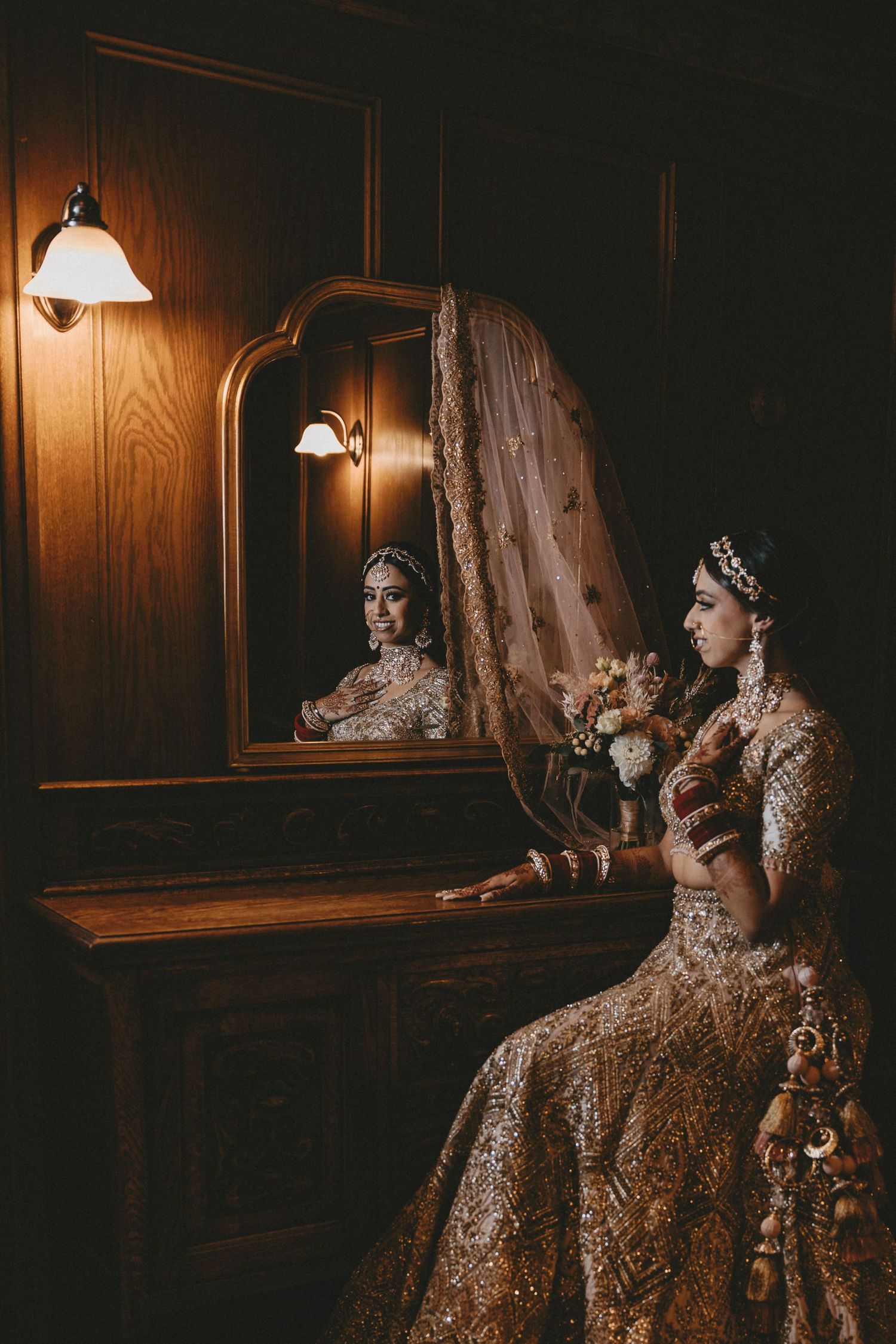 An elegant mirror reflection captures a figure in intricate golden bridal attire with ornate jewelry in a dimly lit room.