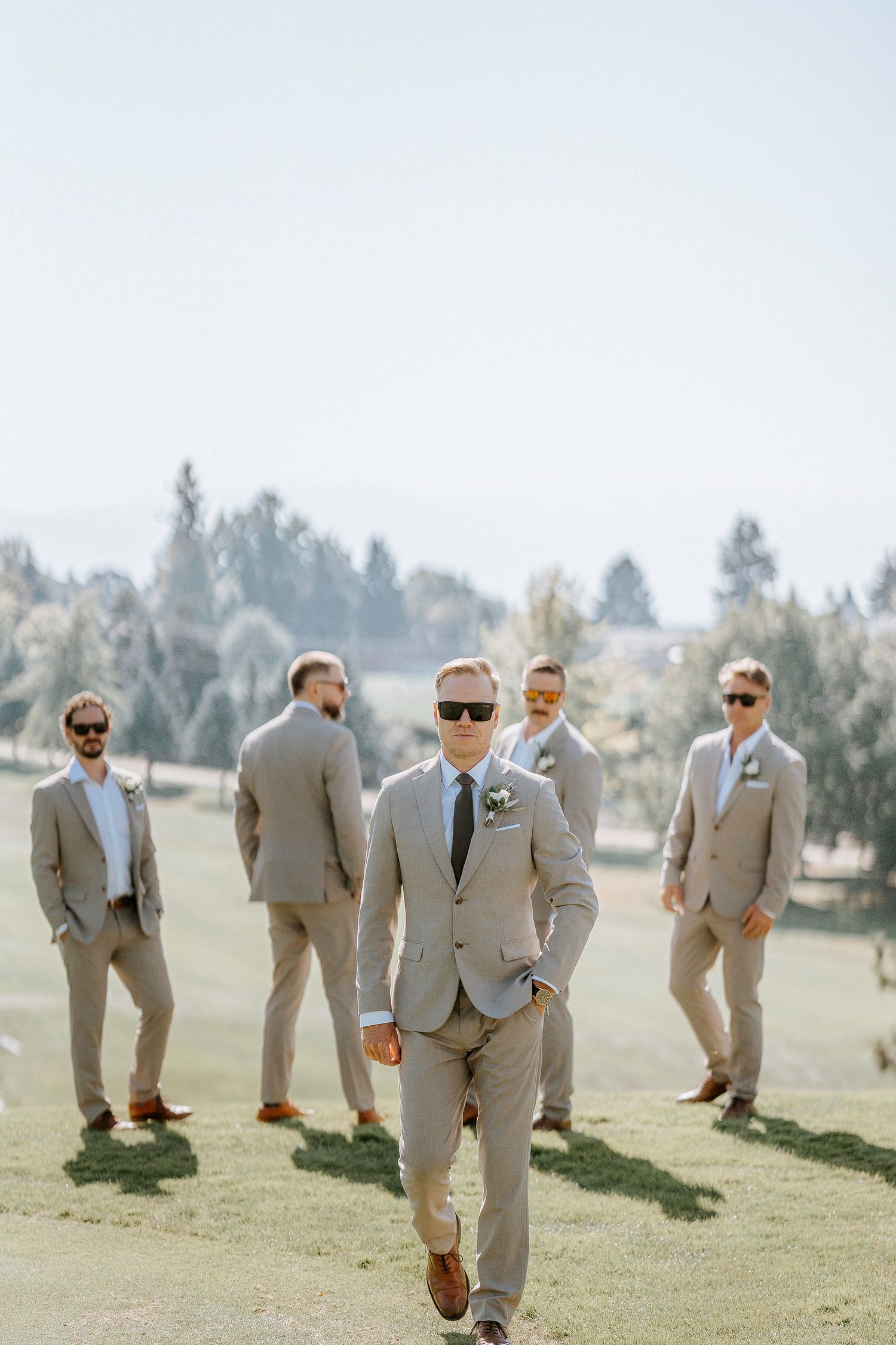 Groomsmen in light gray suits stand together on a grassy hill during an outdoor wedding.