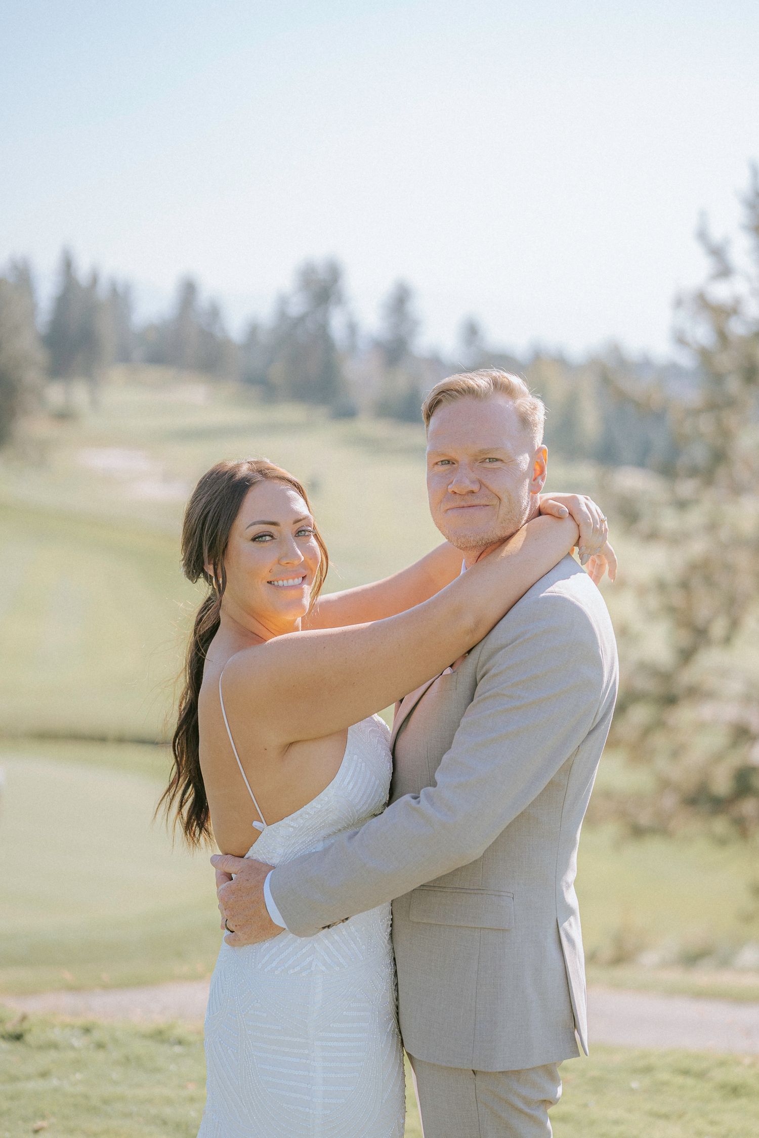 Newlyweds embrace and laugh together during their outdoor wedding photoshoot on a grassy hillside.