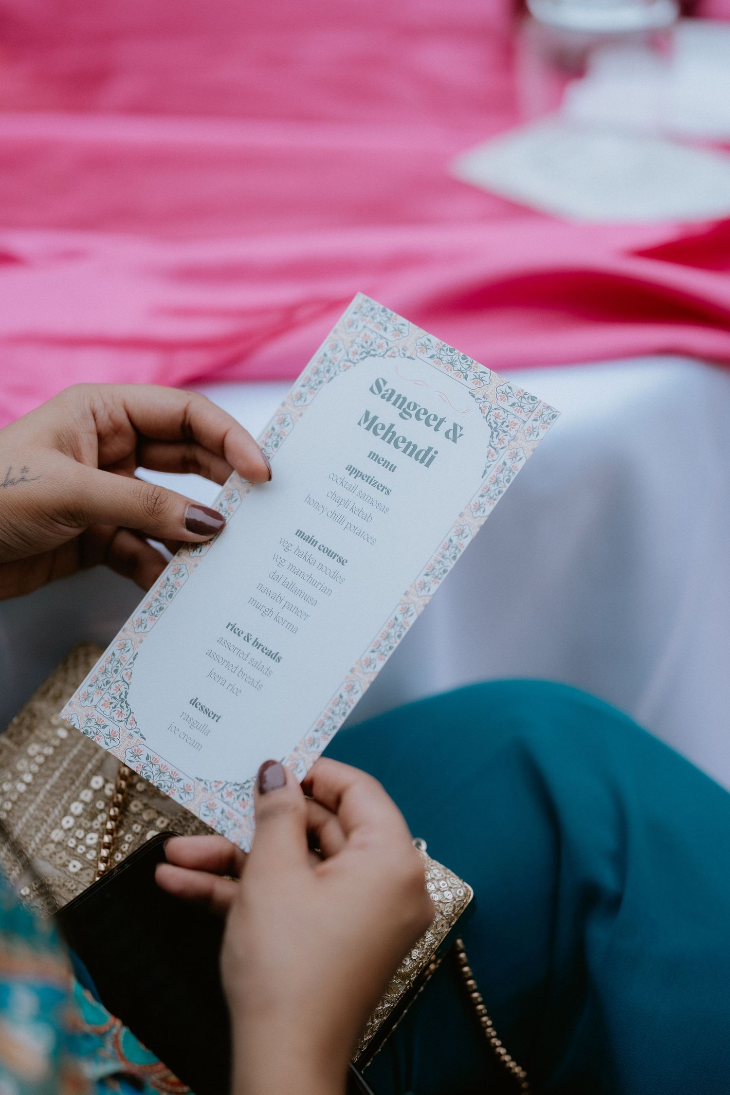 Hands holding an elegant wedding invitation card with delicate lace-like border details against a pink background.