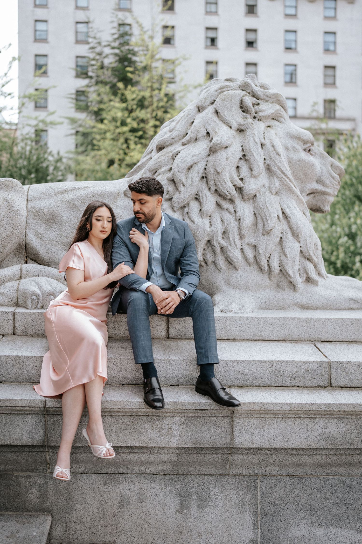 A couple sits together on stone steps next to a decorative lion statue in an urban setting.