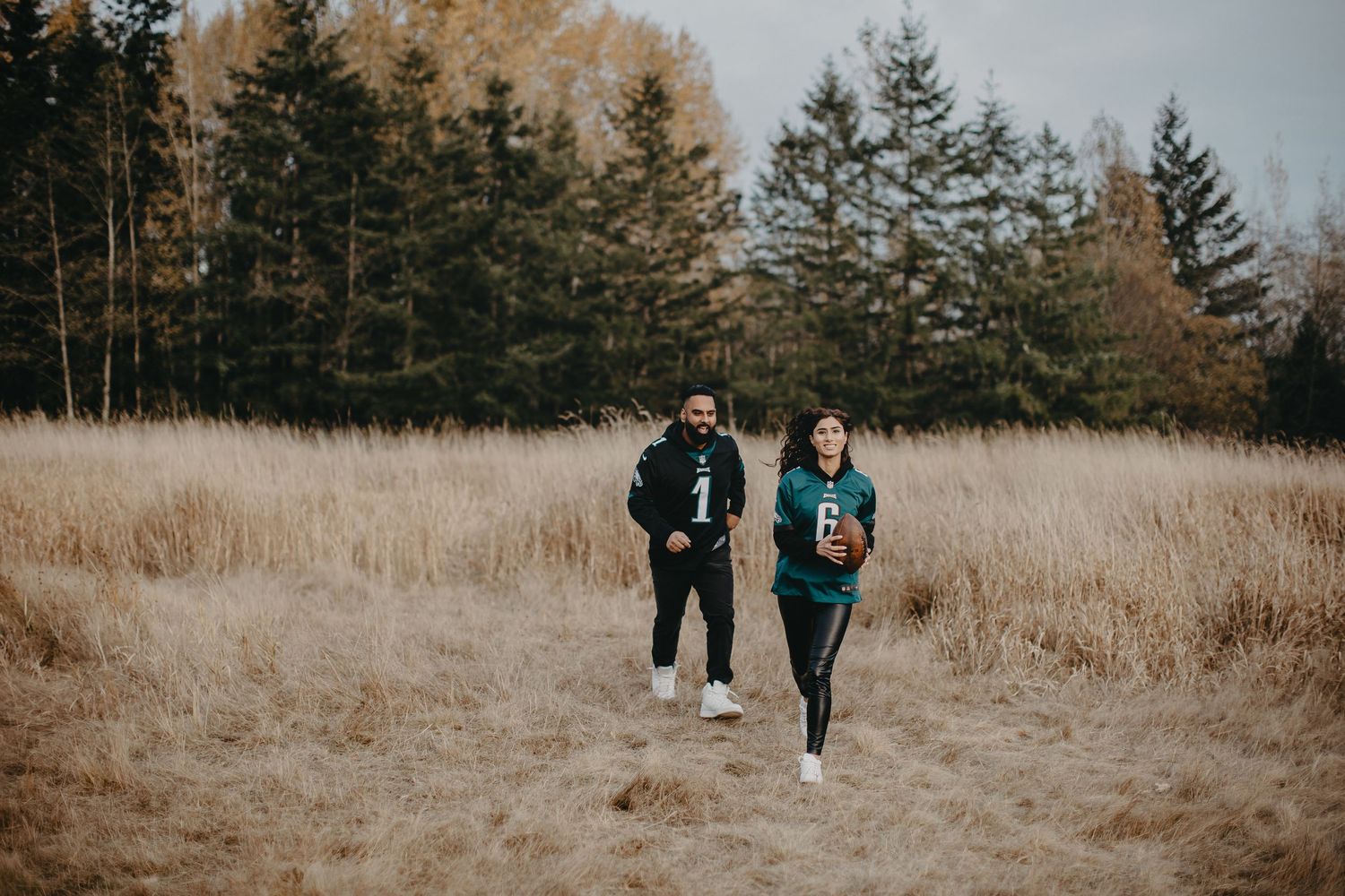 Two people in sports jerseys walking together through tall grass with evergreen forest backdrop.