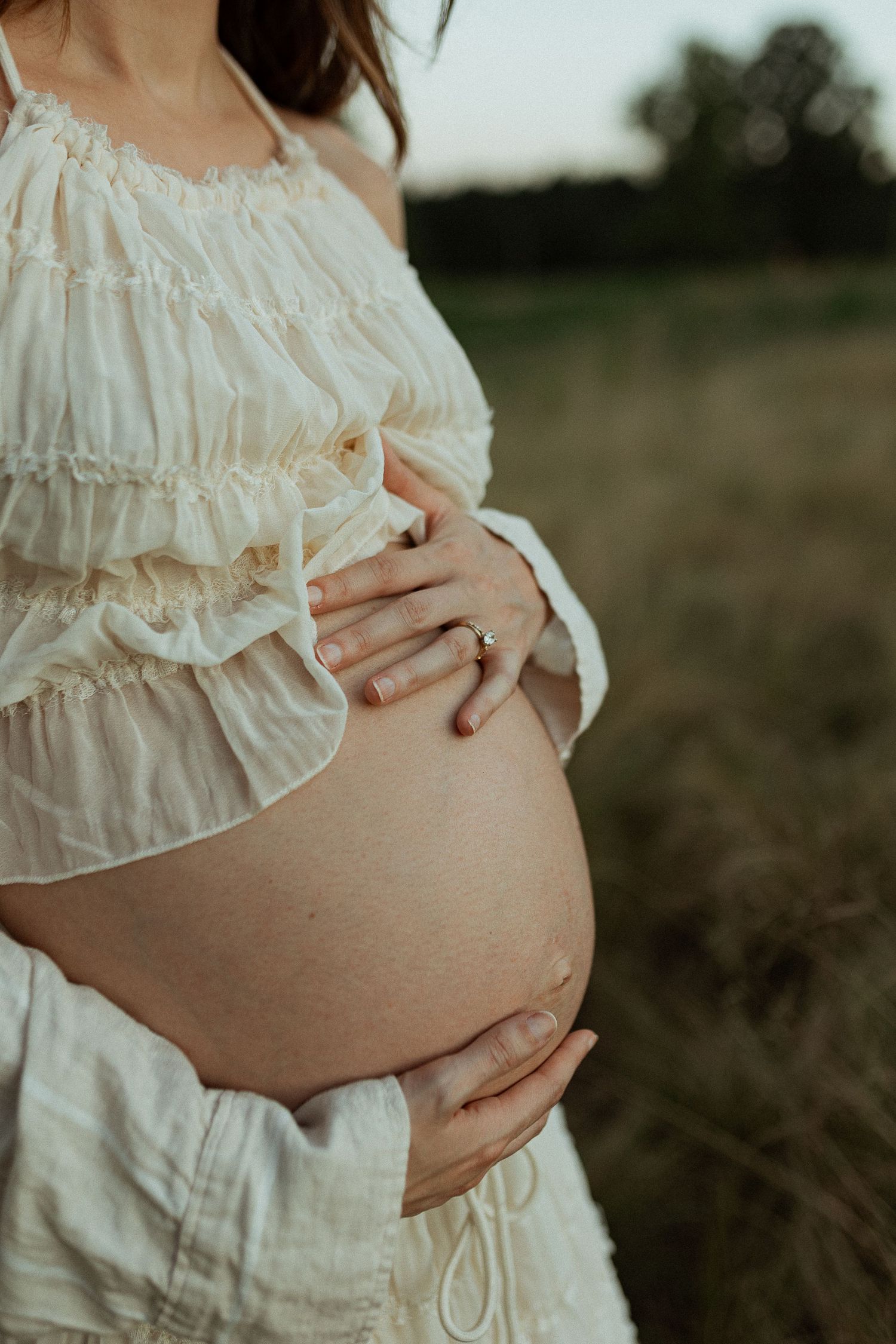 Close-up of pregnant belly in white ruffled dress against natural outdoor backdrop.