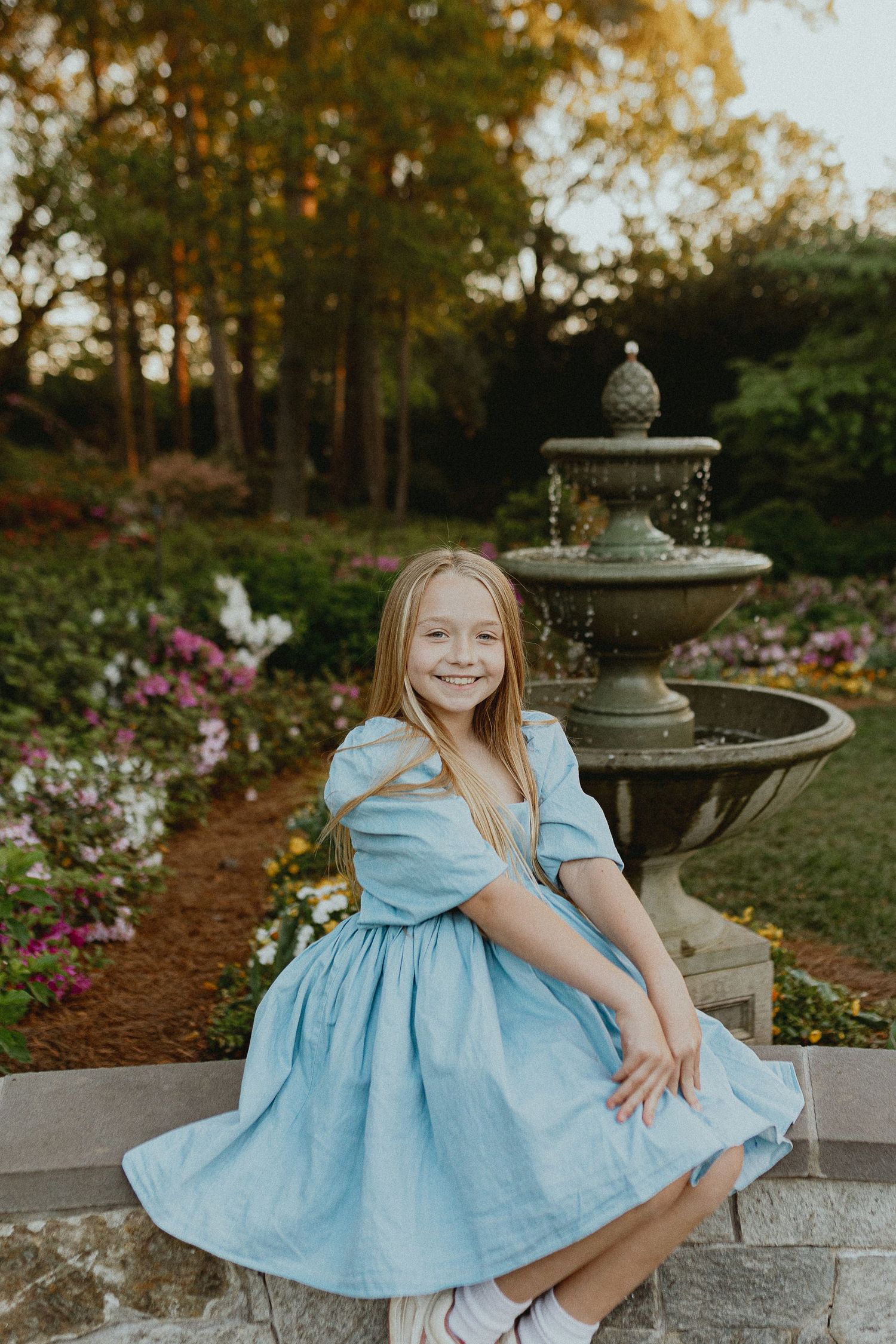 A light blue dress against a garden backdrop with fountain and flowers in warm evening light.