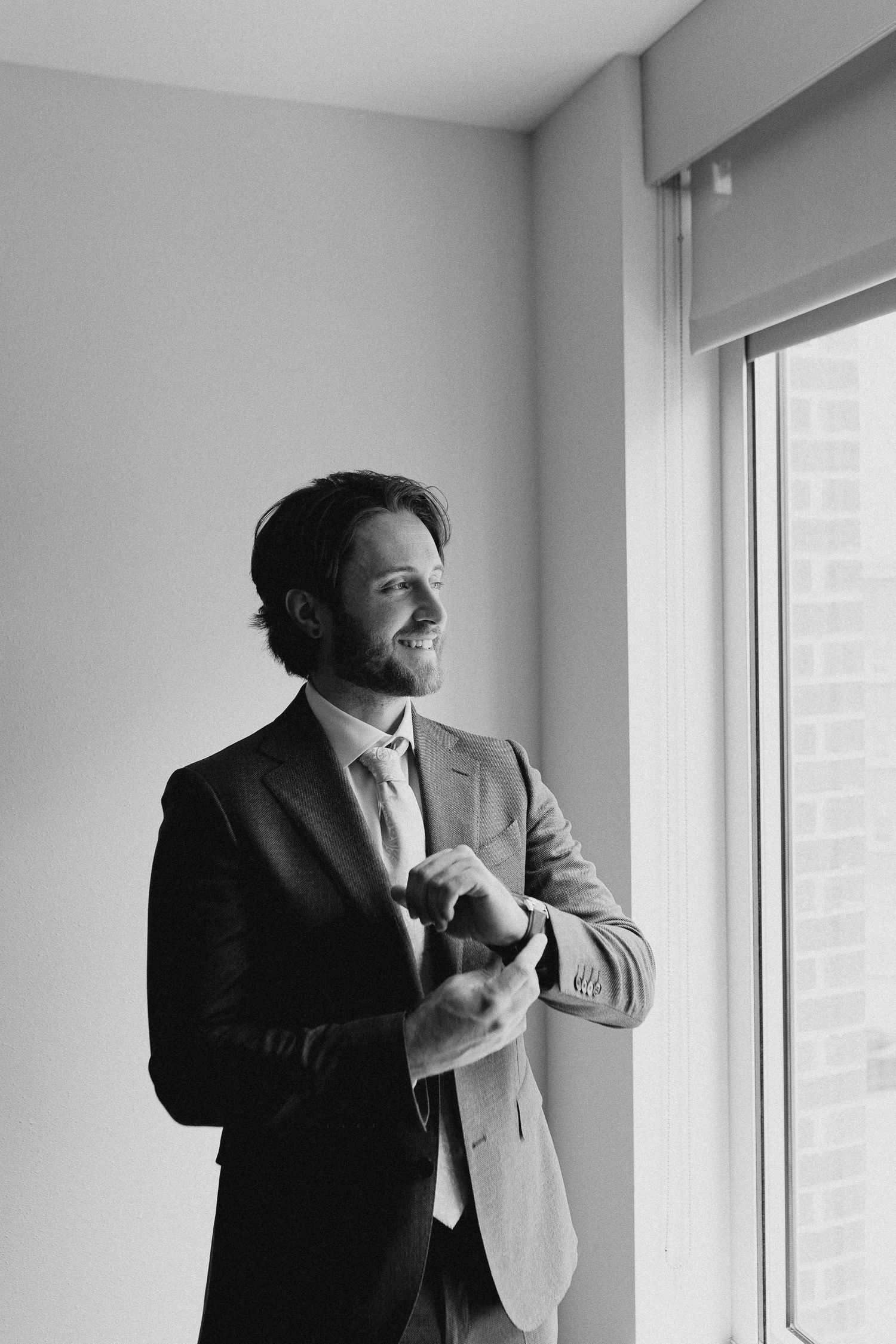 Black and white photo of a smiling groom in a suit adjusting his cufflinks by a window.