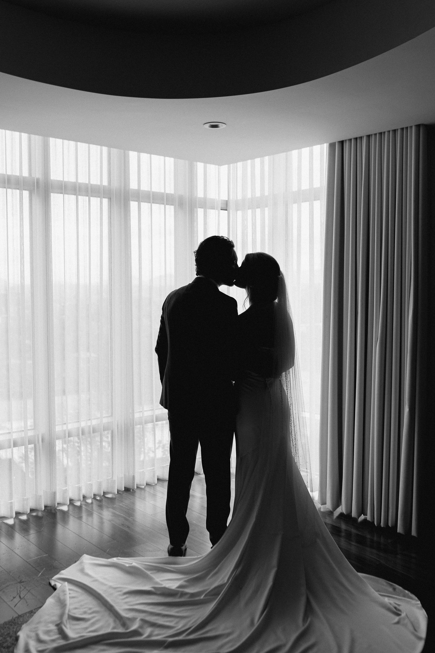 Black and white silhouette of bride and groom kissing before large windows, wedding dress train spread elegantly on floor.