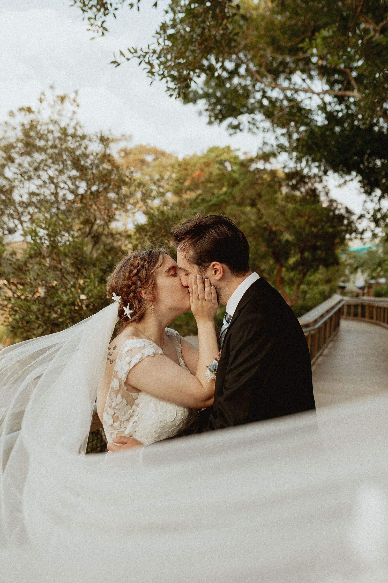 A romantic moment captured on a wooden boardwalk with flowing white veil and natural greenery overhead.