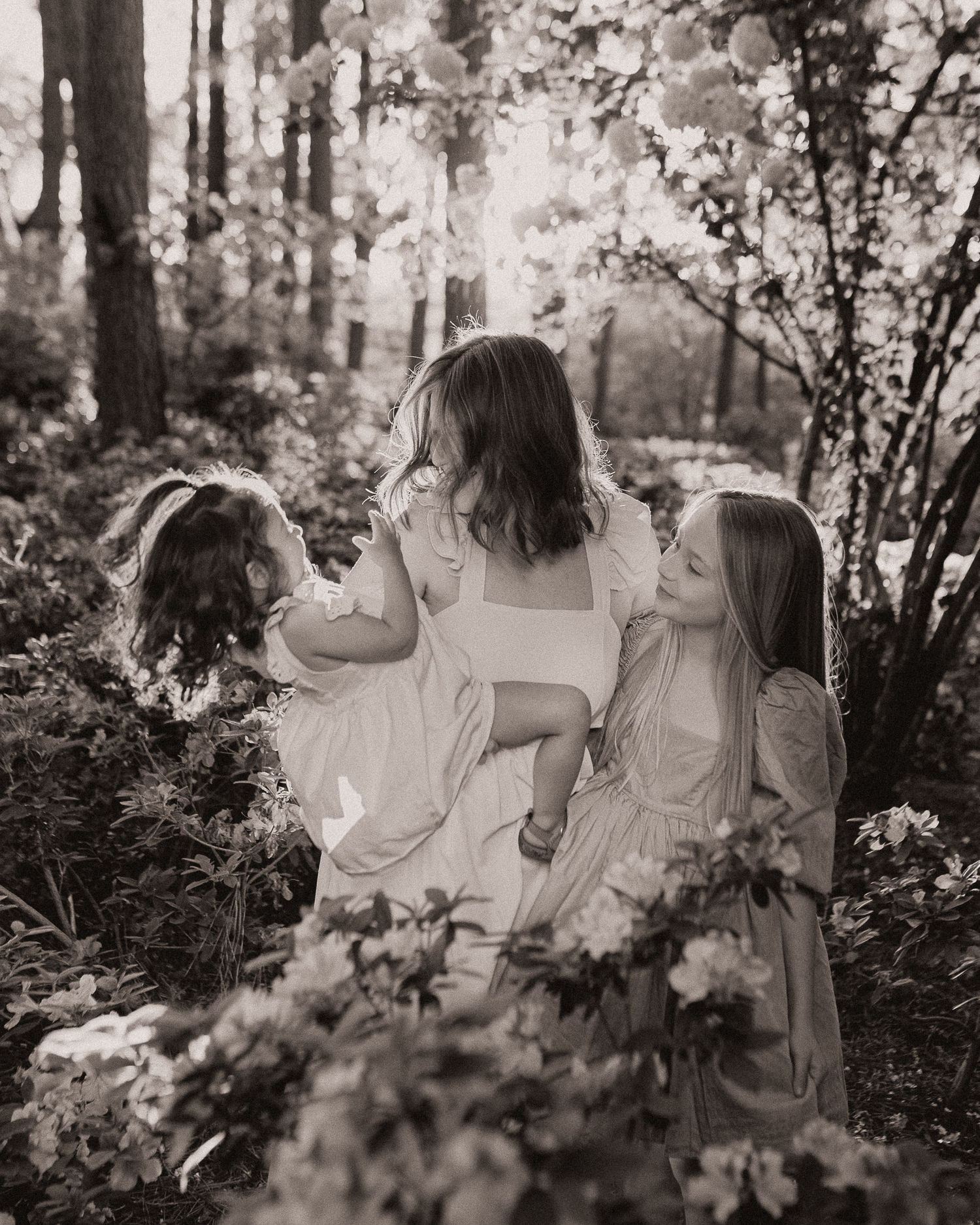 A black and white photo capturing three people in white dresses among flowering bushes.